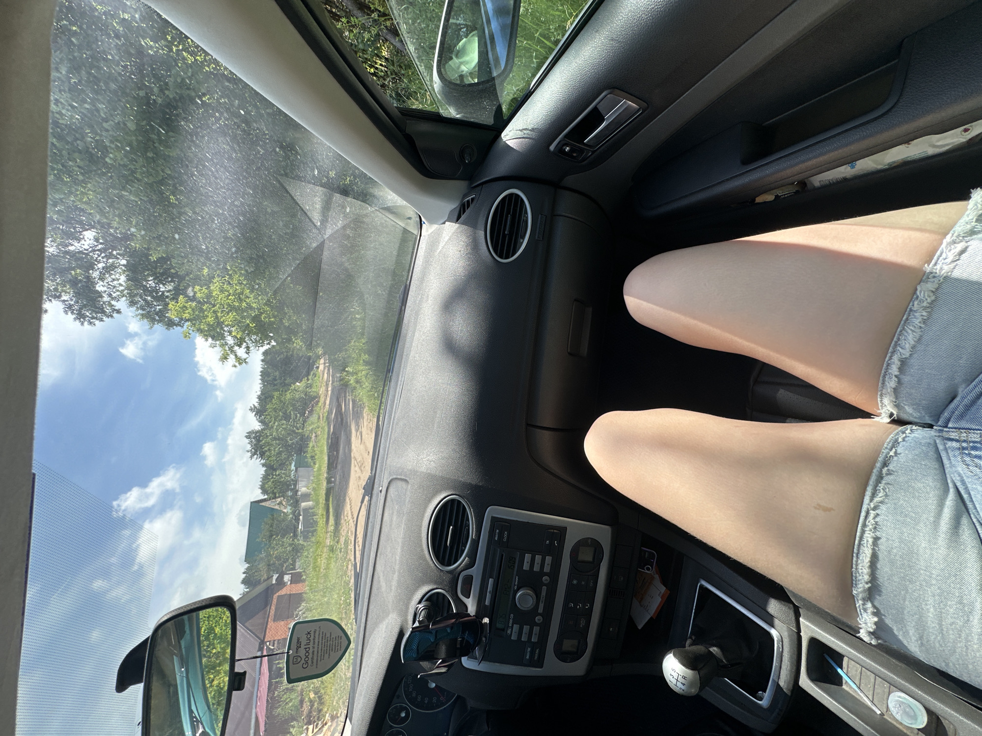 The image is taken from the interior of a car, looking forward through the windshield. A person's legs, wearing denim shorts, are visible in the foreground, suggesting they are the driver or a passenger in the front seat. The dashboard is visible, with the car's stereo and air vents. Outside the car, a dirt road leads through a rural landscape with trees and houses. The sky is blue with scattered clouds. Based on the presence of typical Russian rural houses and vegetation, this scene is consistent with a location in the city of Chaikovsky, Russia.