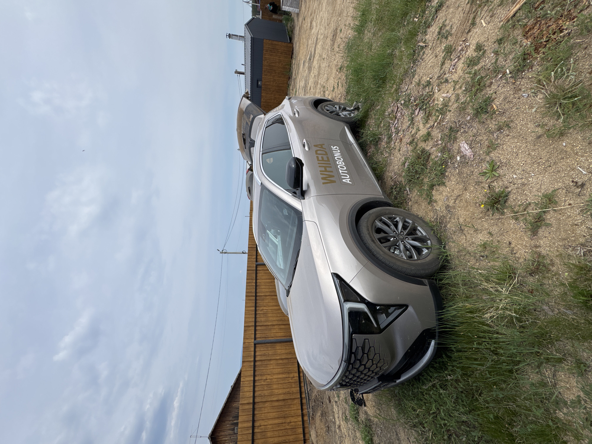 A light-colored SUV is parked on a dirt road with patchy grass. The car's trunk is open. Behind the car, a wooden fence runs horizontally across the image. Further back, there are several small structures, including a dark-colored building with a metal roof and a chimney. The sky is overcast. A power pole with wires is visible to the right of the wooden fence. The scene appears to be in a rural or semi-rural area, consistent with Khuzhir, Russia, known for its natural landscapes and remote setting on Olkhon Island.