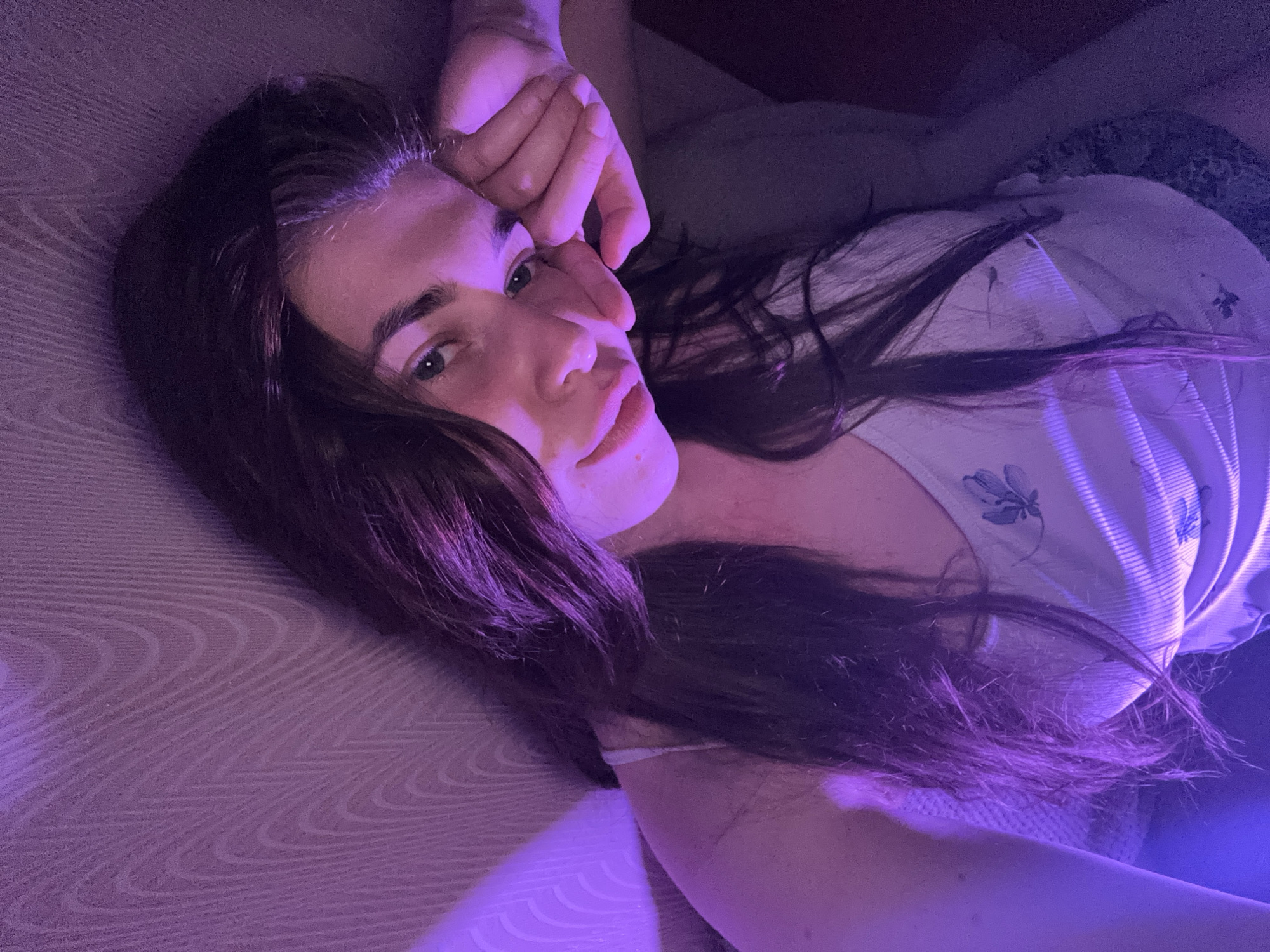 A young woman with dark hair, wearing a white tank top with a floral pattern, is lying down and resting her head on her hand. She is illuminated by purple light, casting a purplish hue on her skin and the textured wall behind her. The woman's face is partially obscured by her hair and hand. The background appears to be an indoor wall with a wood grain pattern. There is no indication of the location being in the city of Мещёры, Russia.