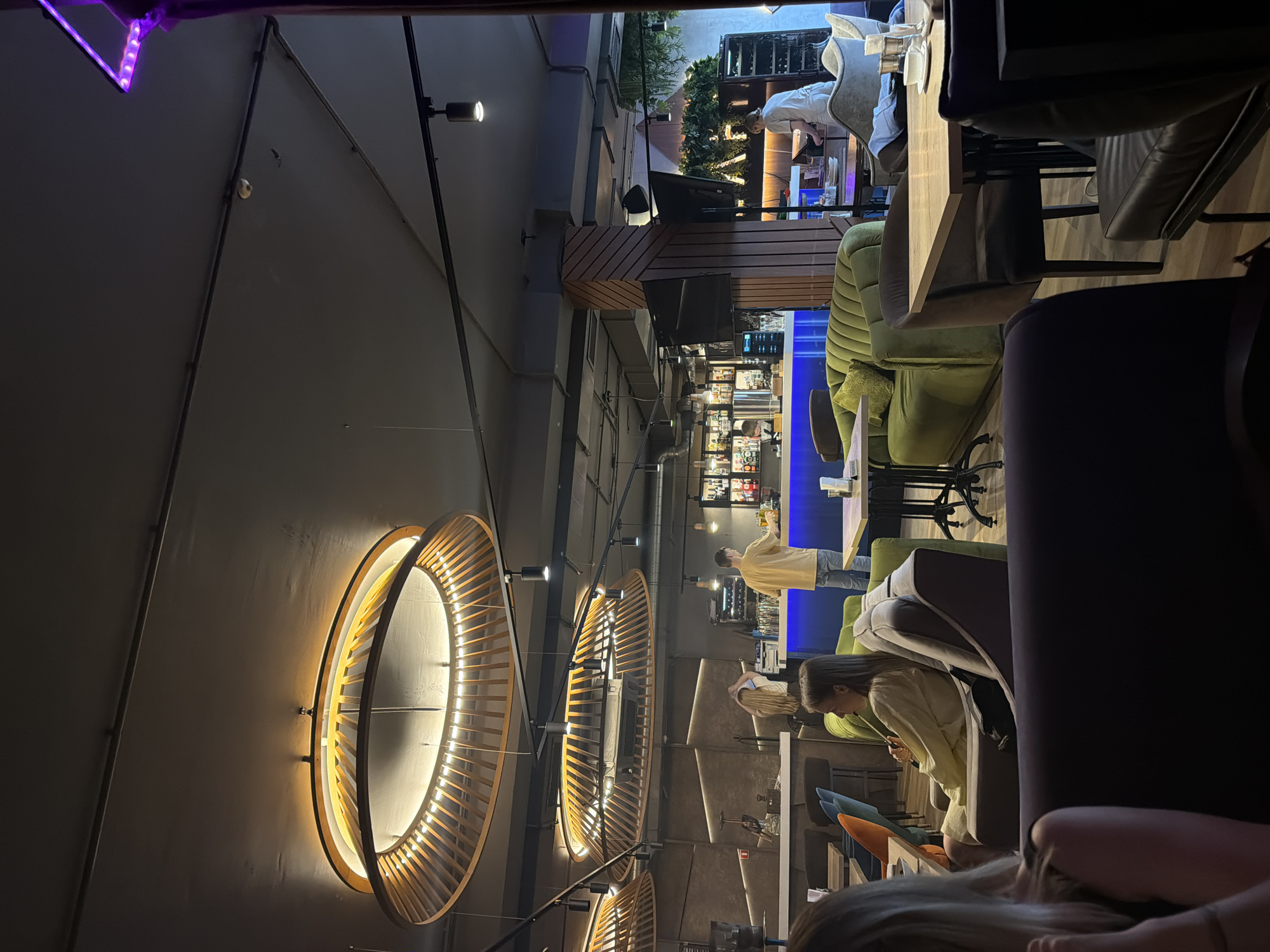 The image depicts the interior of a modern hookah lounge, likely located in Kazan, Russia. The atmosphere is dimly lit, with ambient lighting from distinctive, circular wooden chandeliers and colored LED lights behind the bar.
In the foreground, several patrons are seated. On the left, the back of a woman with blonde hair is visible, partially obscuring the view of the seating area. Further back and to the right, a woman is seated on a plush green sofa, holding a hookah hose to her lips, indicating she is smoking. She is wearing a light-colored top.
Behind the green sofa, a bar area is active. A man in a yellow t-shirt is standing behind the bar, possibly serving or preparing drinks. Another woman, wearing a white uniform, is seen working at a counter to the right of the bar, near a wine rack and what appears to be a music or sound system. Bottles of alcohol are visible on shelves behind the bar.
The furniture consists of comfortable-looking sofas and chairs, with tables placed strategically for patrons. The walls have a textured finish, and decorative wooden elements are incorporated into the architecture and lighting fixtures. The ceiling is dark, with exposed pipes and track lighting. The overall impression is one of a contemporary and relaxed establishment, designed for socializing and enjoying hookah.
