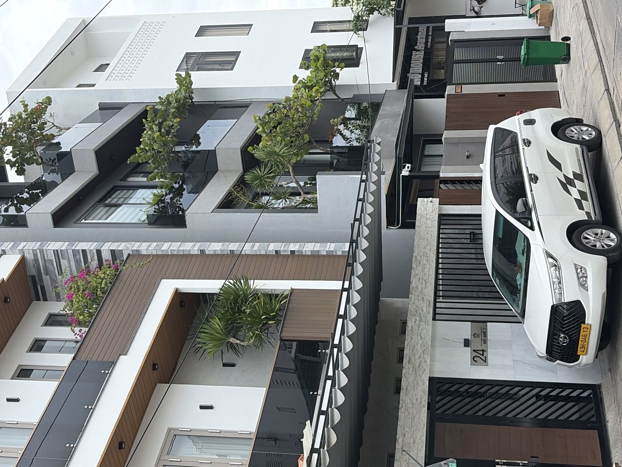 A white Toyota Innova van is parked on the street in front of a modern building with multiple balconies featuring lush greenery. The building has a contemporary design with white, gray, and brown facades, and large windows. The van, license plate 43H-046.13, has a checkered flag design on its side. A green trash bin and cardboard boxes are visible near the building entrance. The street appears to be paved, and the sky is overcast. A sign on the building reads "Mano House Apartment". The location is in Da Nang, Vietnam.