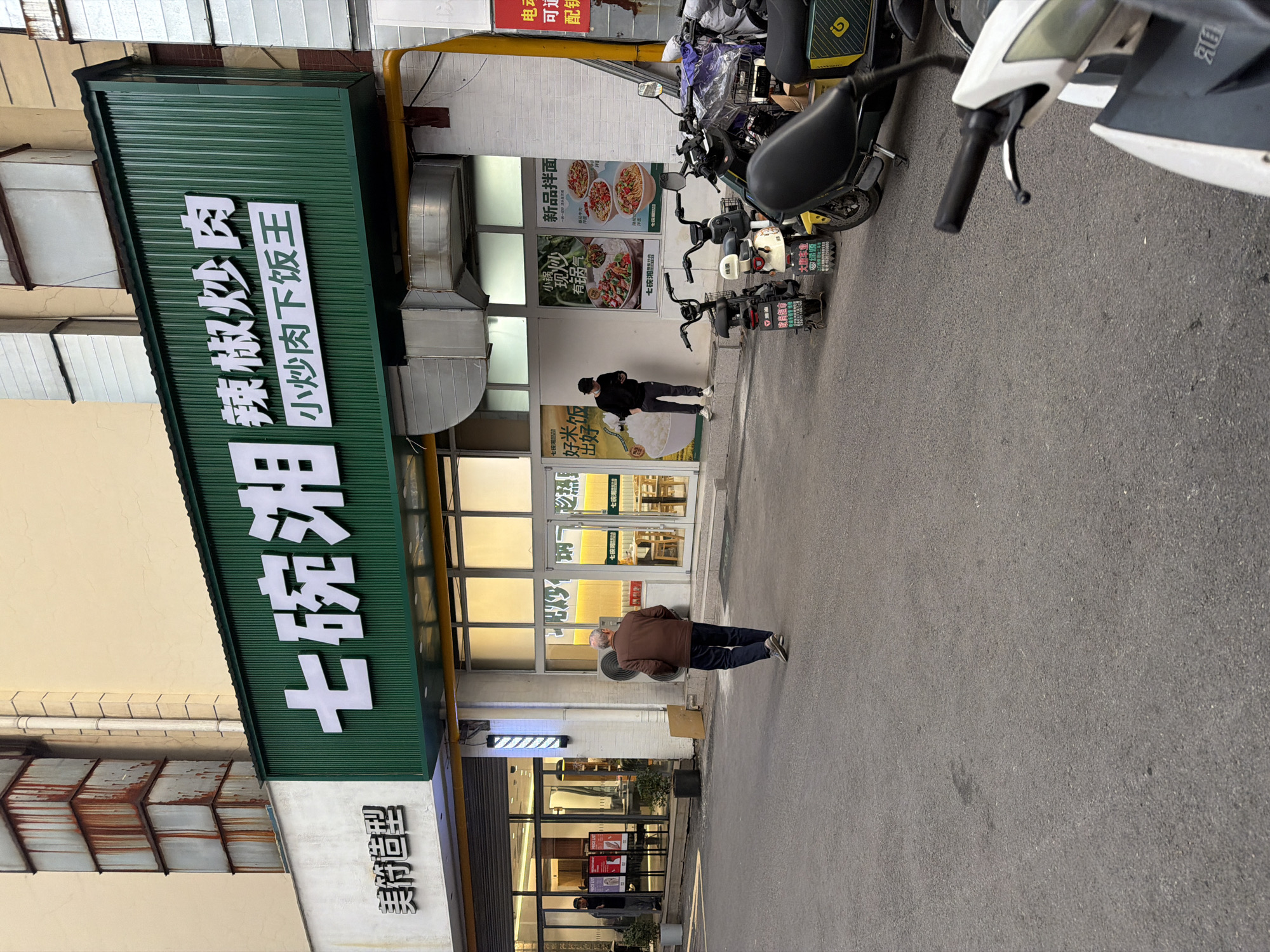 Here is a detailed description of the image:
**Scene:** An exterior shot of a street with shops in Jiangning District, China.
**People:**
* An elderly man in a brown jacket and dark trousers is walking down the street.
* Another person wearing dark clothing is standing in front of a restaurant.
* A third person can be seen through the windows of a shop on the ground floor.
**Objects:**
* Several electric scooters parked on the right side of the street.
* Signs for various businesses including "七碗湘辣椒炒肉" (Qi Wan Xiang Chili Fried Meat).
* Shopfronts with glass windows and doors.
* Restaurant menus and advertisements displayed in the windows.
**Actions:**
* The elderly man is walking.
* The person in front of the restaurant is standing still.
**Interactions:**
* People are in proximity to shops, likely customers or passersby.
**Environment:**
* Street with asphalt pavement.
* Buildings with a mix of modern and older architectural styles.
* Overall urban setting with a mix of businesses.
**Additional details**
*The sign for "七碗湘辣椒炒肉" indicates a Hunan cuisine restaurant specializing in chili fried meat dishes. The Chinese signs "美符造型", possibly referring to a beauty salon, are visible too. There are advertisements for food items like "新品拌面", indicating a noodle dish, and signs advertising "绿源" electric scooters.*
