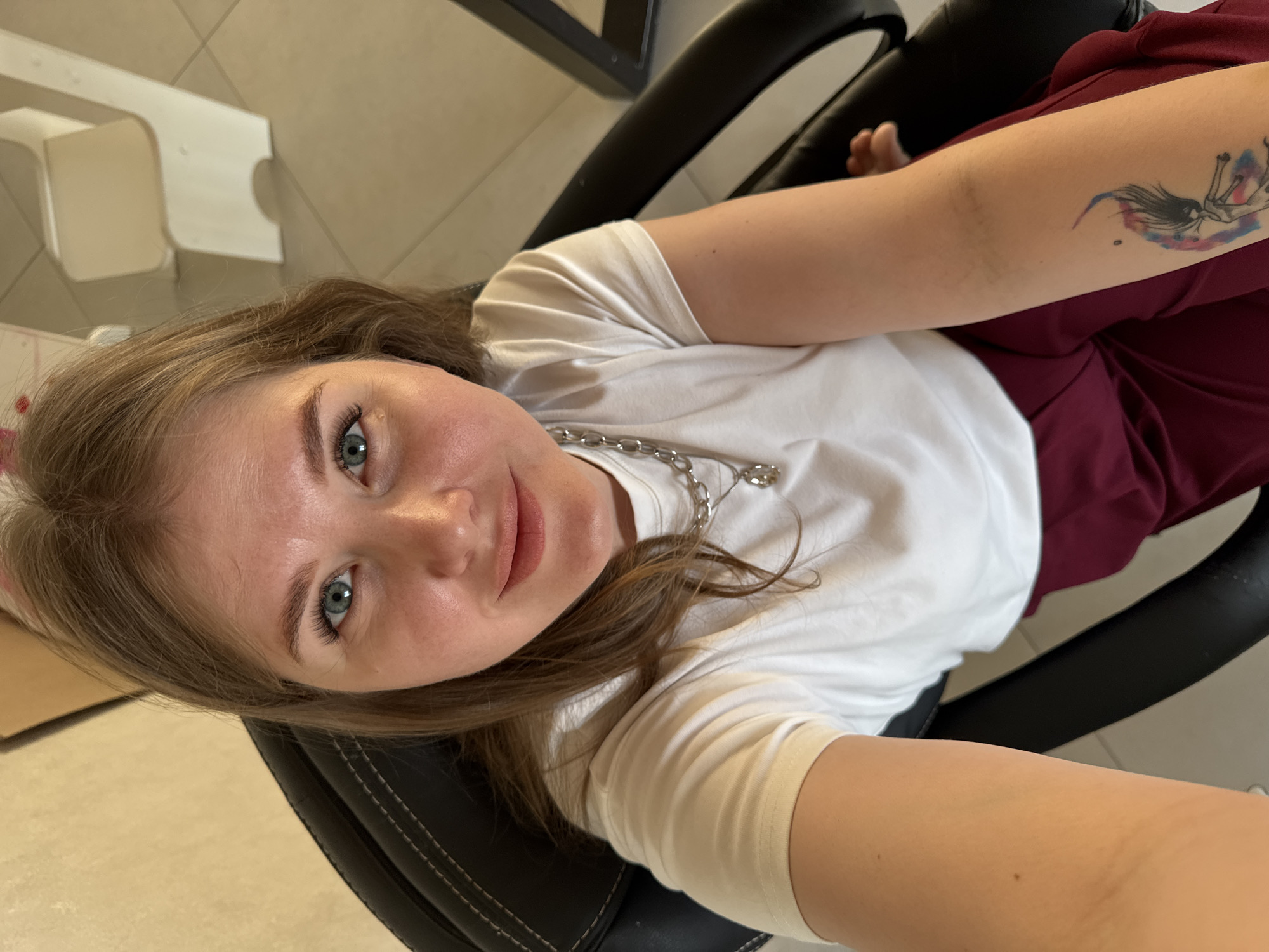 A woman is sitting in a black office chair, facing the camera. She has fair skin, light brown hair, and blue eyes. She is wearing a white t-shirt and a silver chain necklace. On her left arm, there is a tattoo. She is seated in what appears to be an indoor space with light-colored flooring. A white object, possibly a piece of furniture, is visible in the background. The location may be Gornyi (Dvurechenskoe S/P), Russia.