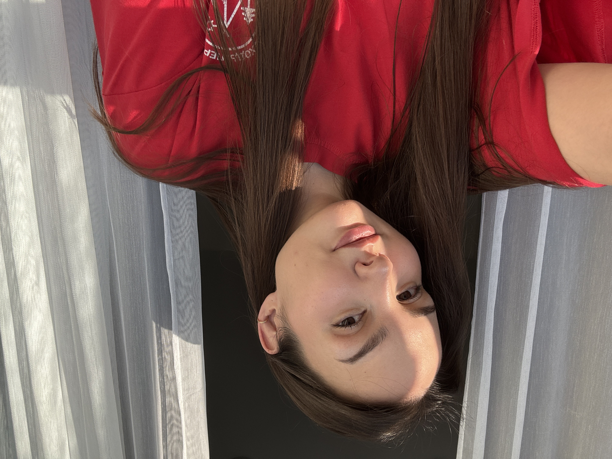 A young woman with long brown hair is posing for a photo. She is wearing a red t-shirt with white lettering. The photo appears to have been taken indoors near a window with white, semi-transparent curtains. The location is not apparent from the image and therefore cannot be confirmed as Chaikovsky, Russia. The woman is looking at the camera with a slight smile.