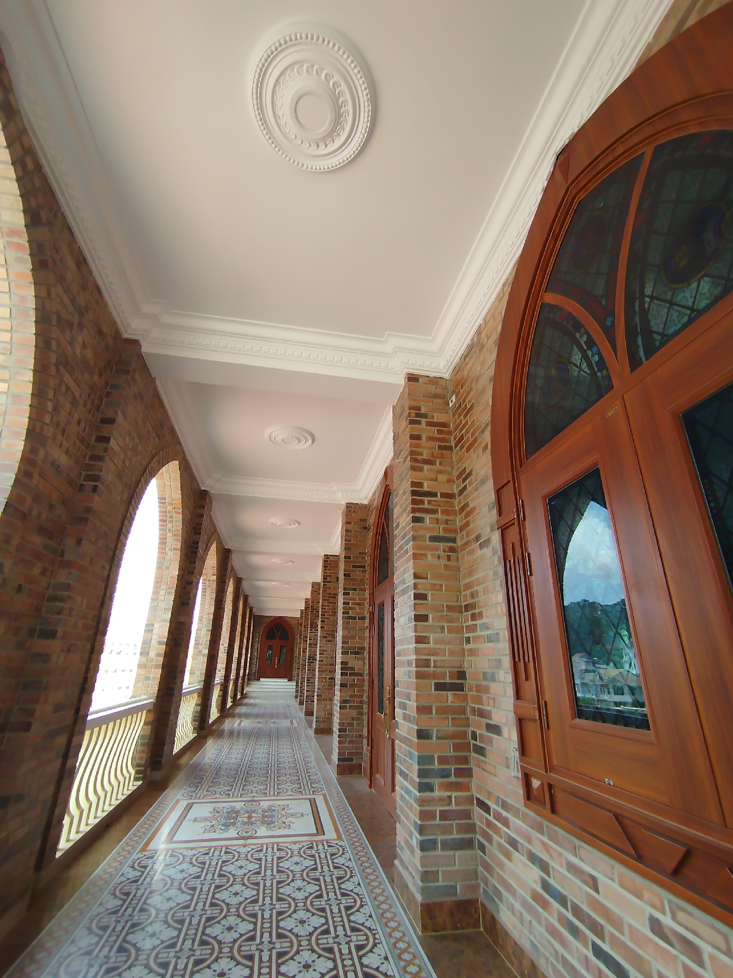 The image captures a long, covered corridor in a building likely located in Cam Ly, Đà Lạt, Vietnam. The corridor features a series of arched openings on one side, providing views to the outside, where a white building and hillside foliage are visible. The opposite side of the corridor is lined with large arched windows or doors made of dark wood and containing stained glass or clear panes reflecting the external environment. The walls are constructed of multicolored bricks. The floor is covered with patterned tiles in neutral tones, creating a visual pathway down the corridor. The ceiling is white with decorative molding and circular medallions spaced along its length. The perspective suggests the photograph was taken at a low angle, emphasizing the corridor's depth. No people are visible in the immediate foreground.