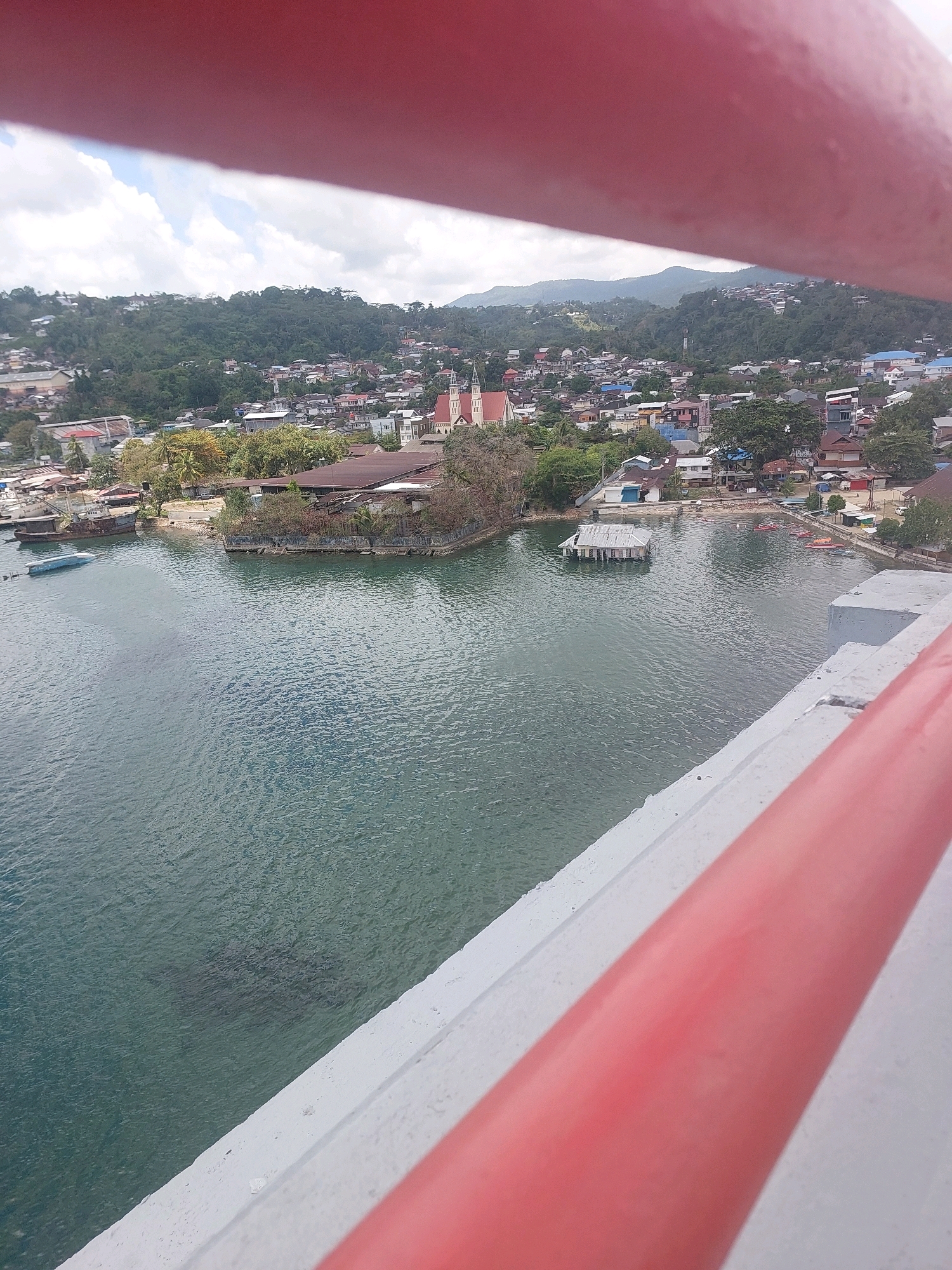 This image shows a view of a coastal city, likely Ambon, Indonesia, from an elevated vantage point. The foreground is dominated by a red and white railing, part of a bridge or elevated walkway. The middle ground features a bay with calm, greenish water. Several boats are visible, including a large, old-looking barge with a rusty hull, and a smaller blue boat partially submerged. In the bay, a floating structure with a corrugated metal roof stands out.
The background reveals a densely populated urban area built on a hillside. Buildings of various sizes and colors are clustered together, with many roofs visible. Lush green vegetation covers the slopes behind the city. Prominent structures include a church with a steeple and a red-tiled roof, and other commercial or residential buildings. The overall scene suggests a vibrant coastal community. The sky is partly cloudy, indicating daytime. The specific location appears to be a harbor or bay area within Ambon city, possibly overlooking the Poka area or the vicinity of the sea.