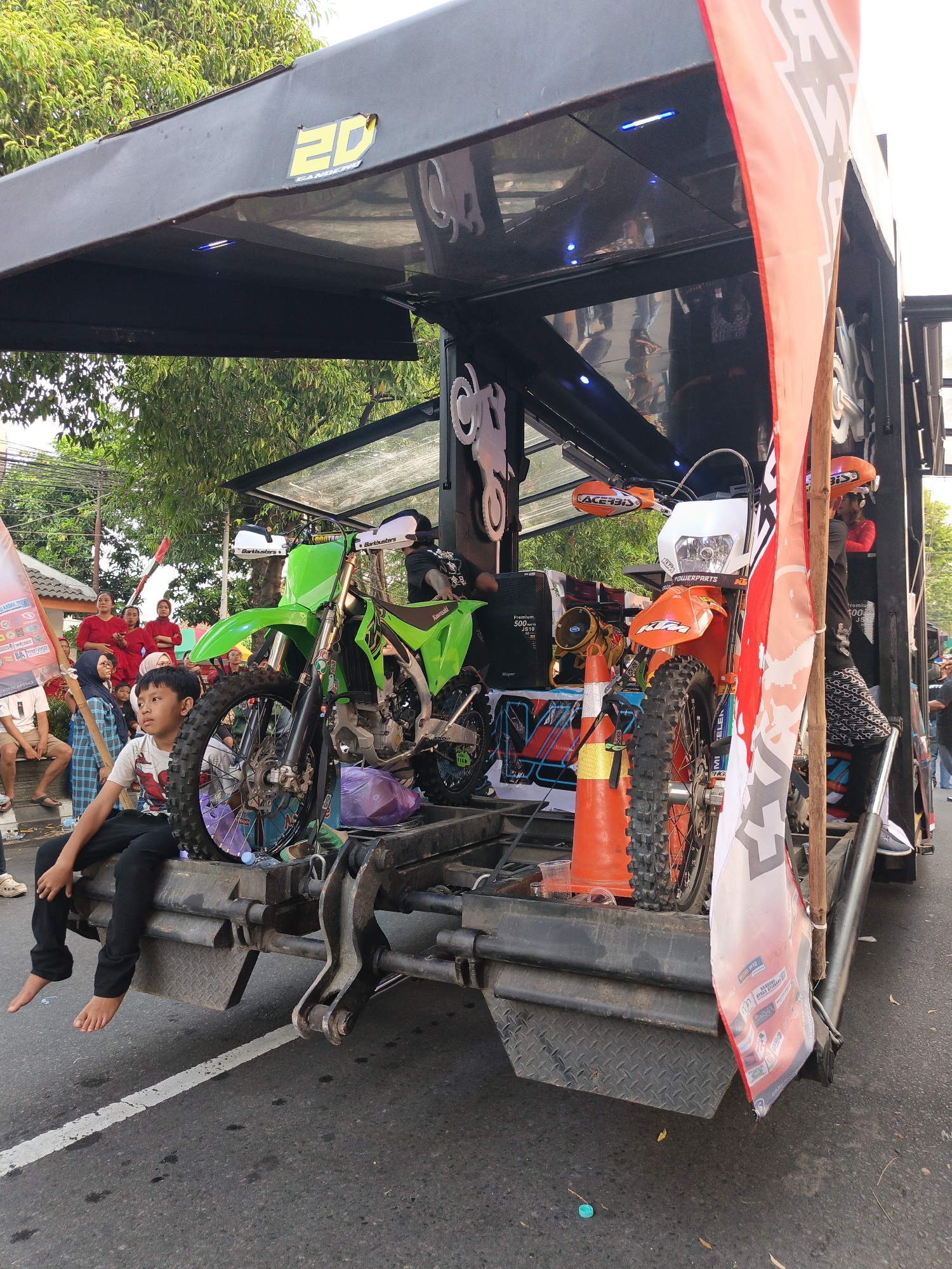 In Gayamprit, Indonesia, a boy sits on the back of a black flatbed truck, his legs dangling over the side. The truck is loaded with two dirt bikes: one bright green and the other orange. The green bike is positioned on the left, and the orange bike on the right. Both bikes are facing forward, their handlebars and front wheels clearly visible. A large, red and white flag, partially unfurled, hangs from the right side of the truck, obscuring some of the vehicle's details.

Behind the dirt bikes, the interior of the truck's structure is visible. There are blue LED lights illuminating the space, and various logos and signs are displayed. The people in the background, dressed in red and blue clothing, appear to be spectators or participants in an event. The ground beneath the truck is asphalt, with a white line marking a lane. The scene is set outdoors, with trees visible in the upper left corner, suggesting it's daytime.