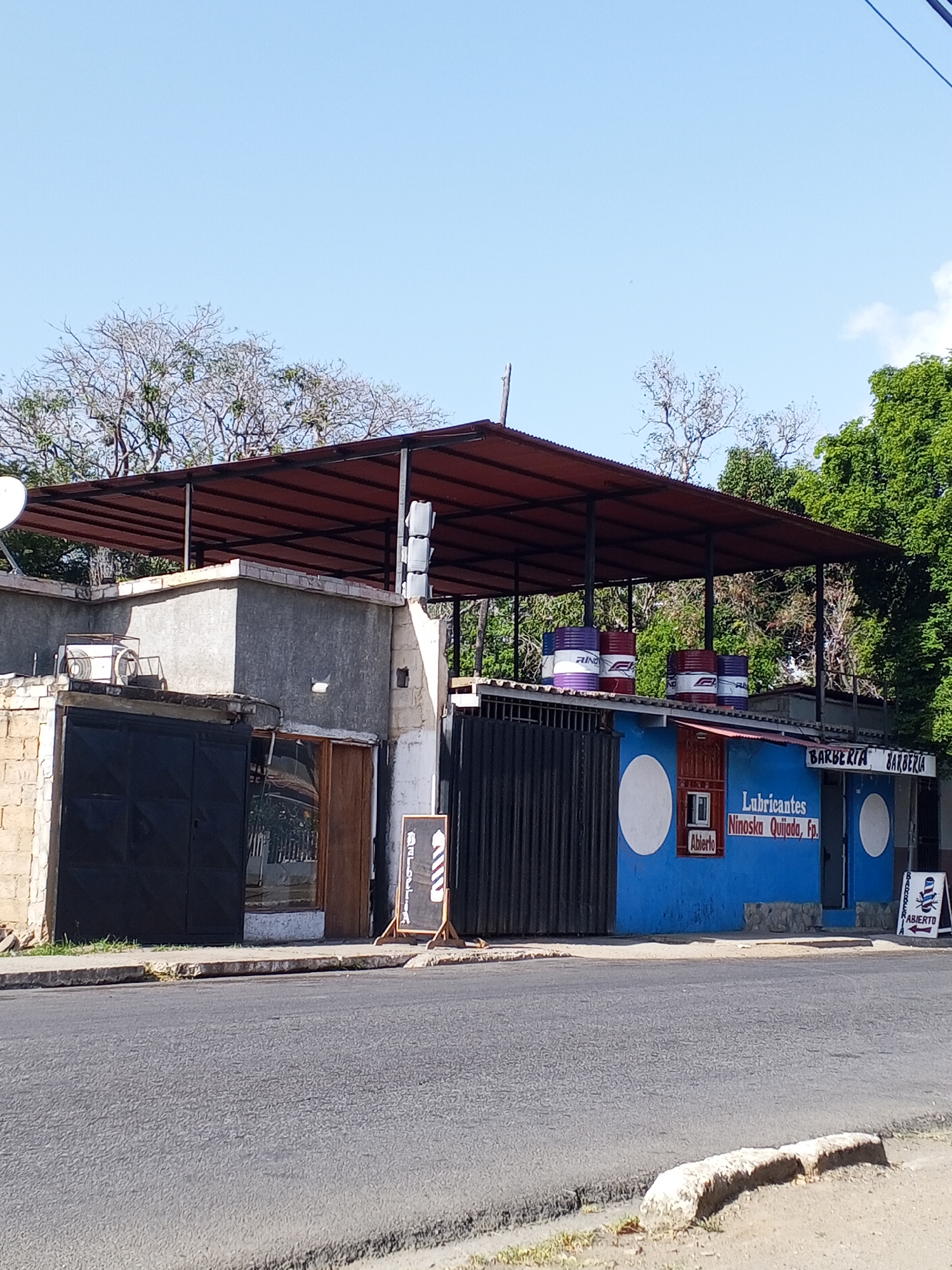 The image depicts a commercial building with a mix of businesses in an urban setting. The building is partially blue and has a sign that reads "Lubricantes Ninoska Quijada, Fp." as well as "BARBERIA BARBERIA" above an entrance. There are also barrels with branding on the roof. To the left of the blue structure is a grey building with a black metal door and a wooden door. There is an overhanging roof supported by metal poles. A barber shop sign is placed on the sidewalk in front of the building. The street is asphalt and appears to be a quiet road. Trees are visible behind the building, and the sky is clear and blue. The location is Unknown, Unknown. No people are visible in the image.