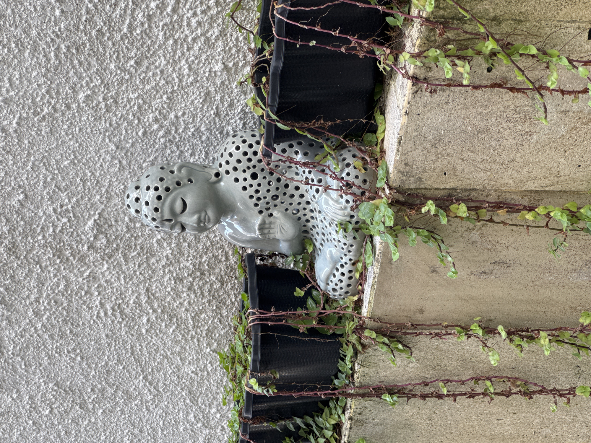 A gray ceramic statue of a seated Buddha with many holes is positioned near a black planter on a wall, with vines and foliage climbing nearby. The statue is set against a textured white wall, while the planter sits adjacent to a concrete wall section. The location is Kuta Selatan, Indonesia.