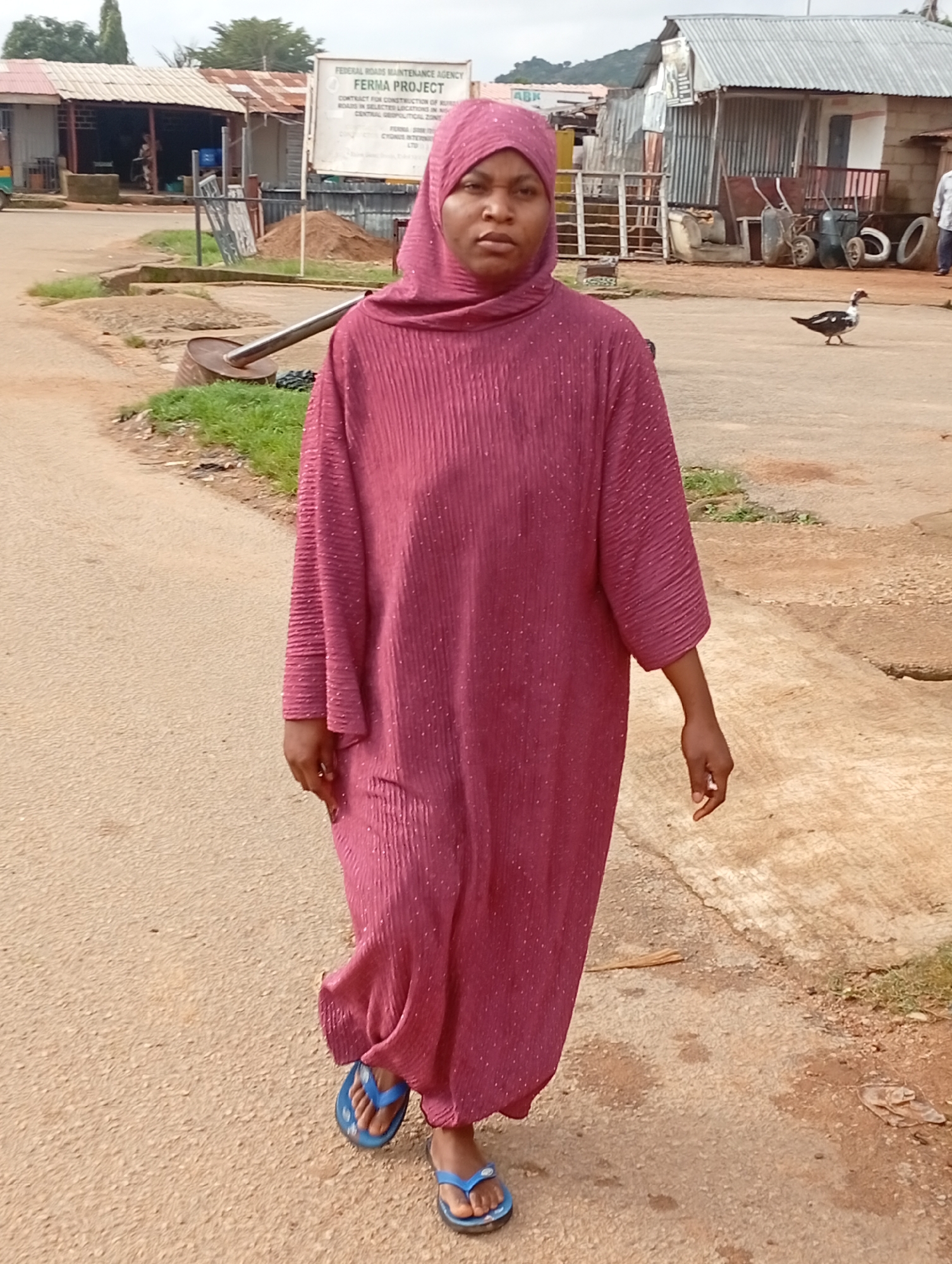 A woman wearing a maroon, floor-length, pleated dress and a maroon hijab walks forward on a dirt road in Jos, Nigeria. She is facing the camera and has her hands at her sides. She is wearing blue flip-flops. Behind her, to the left, are a few small buildings with corrugated metal roofs. A sign that reads "FEDERAL ROADS MAINTENANCE AGENCY FERMA PROJECT" is visible. To the right of the woman, in the background, is a dark-colored duck with white spots on its chest. There are also several metal wheelbarrows and stacks of materials. The ground is a mix of dirt and some patchy grass.