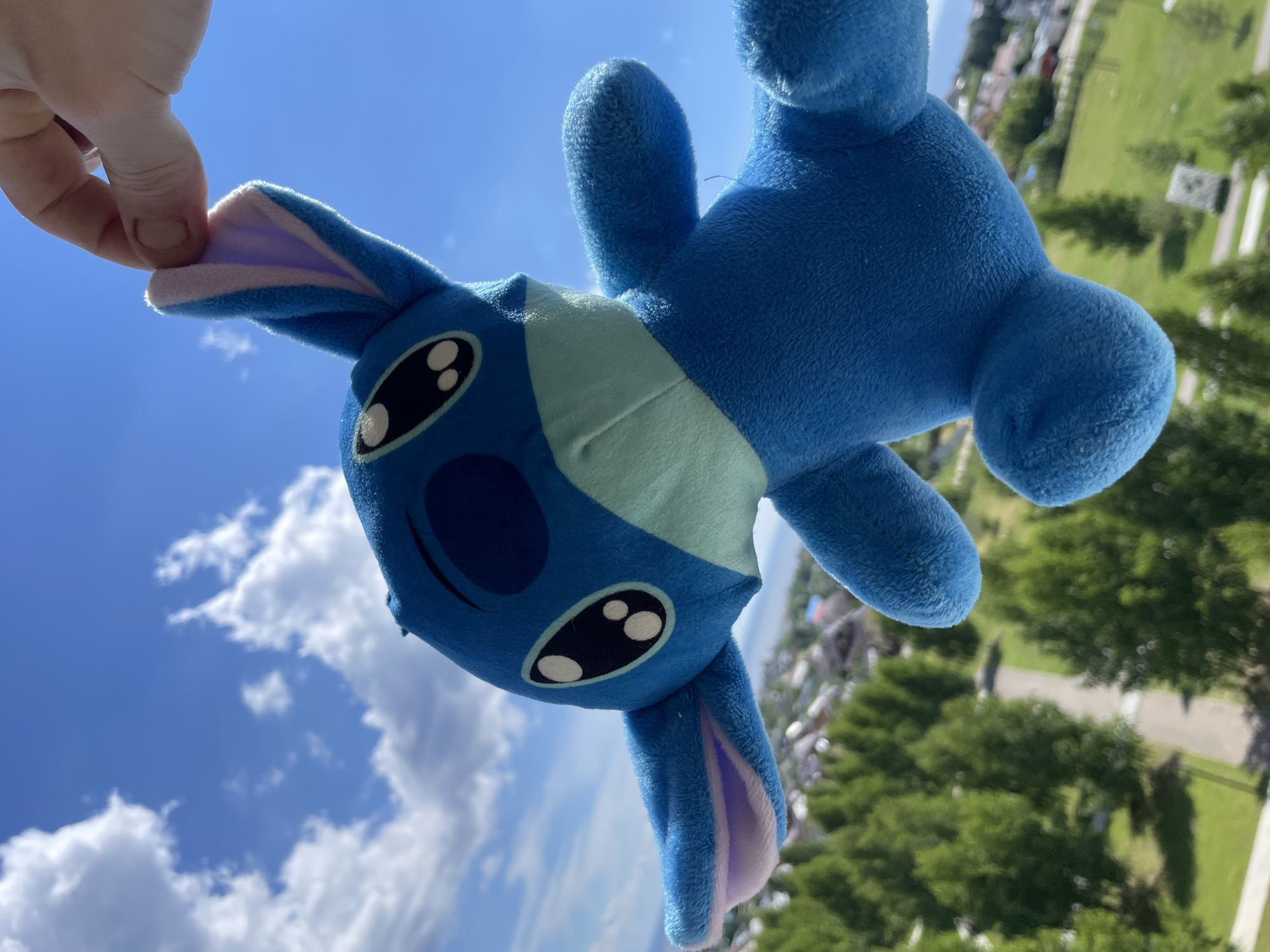 A blue plush toy, resembling the character Stitch, is held up by a human hand against a bright blue sky with scattered white clouds. The toy is positioned in the foreground, with its head tilted slightly upwards. Green trees and a few houses are visible in the blurred background, suggesting an outdoor setting, possibly on the outskirts of a residential area. The location is identified as Laishevo, Russia.