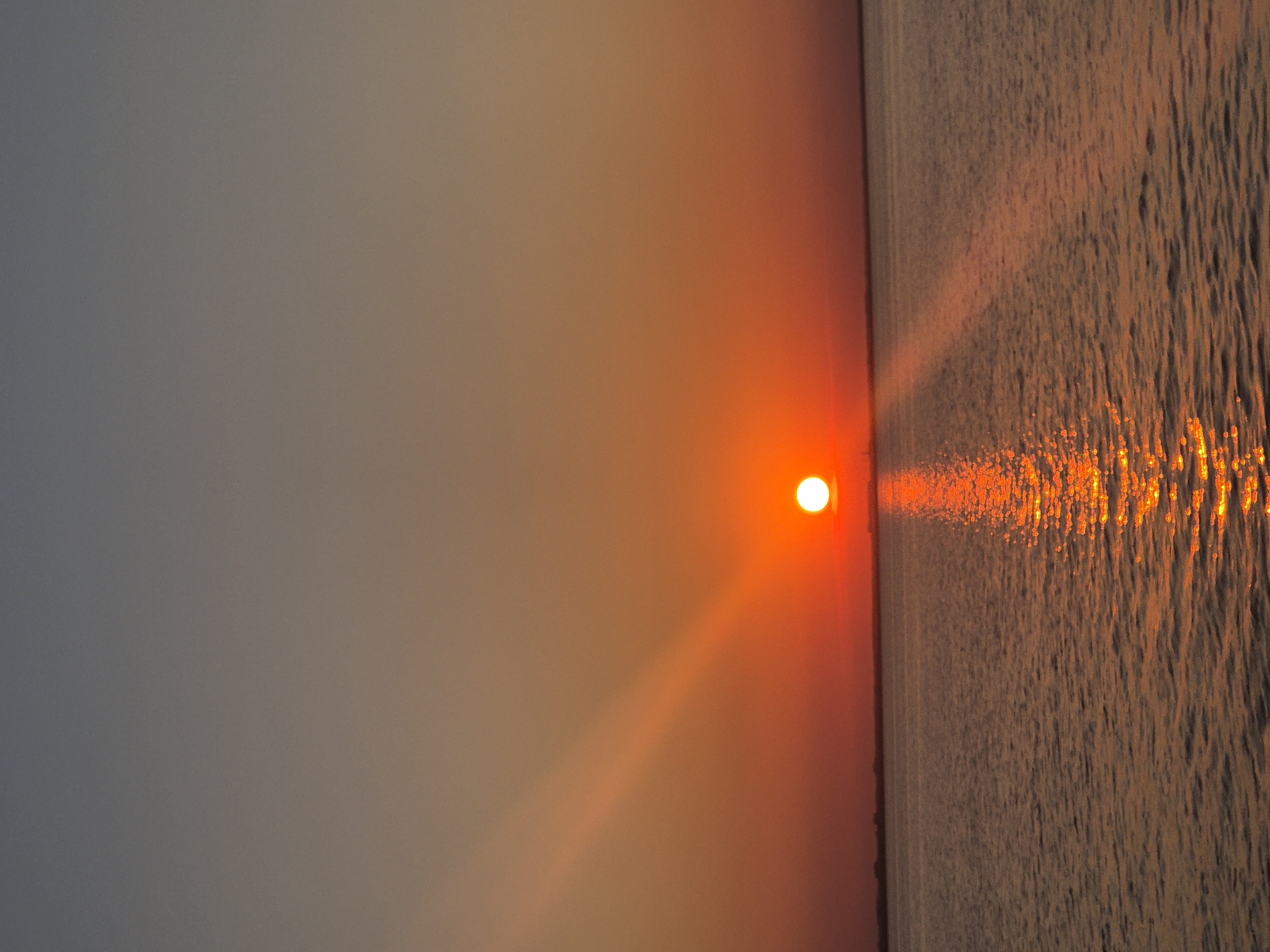The image depicts a serene sunset over a body of water, likely a lake or sea, in Kokshetau, Kazakhstan. The sun, a vibrant orange orb, is positioned just above the horizon, casting a warm, golden glow across the scene. A prominent reflection of the sun shimmers on the rippling surface of the water, creating a pathway of light that stretches towards the viewer.
The sky transitions from a pale grey at the top of the frame to a rich gradient of orange and red closer to the horizon, illuminated by the setting sun. The water's surface is textured with gentle waves, catching the light and creating a dynamic visual effect.
The scene evokes a sense of peace and tranquility, characteristic of a sunset. The warm colors and the gentle movement of the water contribute to a calm and contemplative atmosphere. There are no people or specific objects visible, emphasizing the natural beauty of the landscape. The time of day is clearly evening, as indicated by the setting sun and the warm hues in the sky and on the water. The weather appears clear, allowing for an unobstructed view of the sunset. No text is visible in the image.