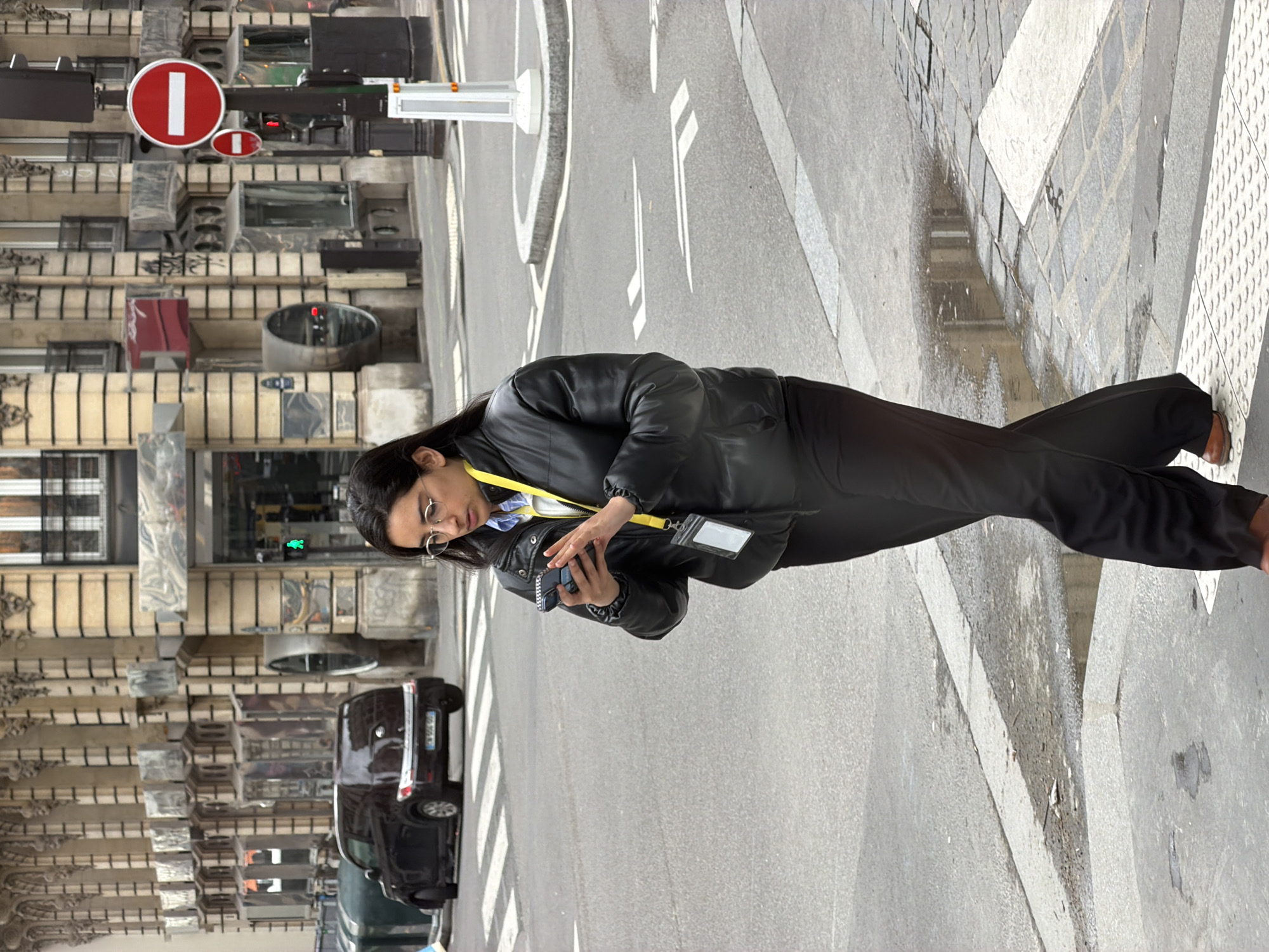 A person wearing a black jacket and pants is standing on a cobblestone sidewalk next to a street in Paris, France. The person is looking at a small electronic device. In the background, there is a street with cars, buildings with ornate facades, and traffic signs. A no-entry sign is visible above the buildings. The street is wet in places, and white lines mark the road.