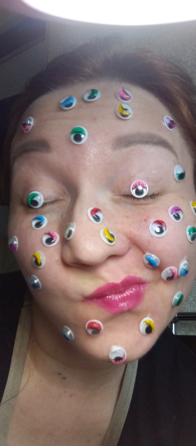 The image displays a close-up shot of a person's face, likely a woman, adorned with numerous colorful googly eyes stuck across her forehead, cheeks, nose, and chin. Her natural eyes are closed, and her lips are pursed, enhanced with bright, glossy pink lipstick. The googly eyes come in various colors, including green, pink, blue, yellow, red, black, and silver, giving the face a whimsical and humorous appearance. Her hair, appearing to be a reddish-brown, is visible framing her face. The background is indistinct and appears to be an indoor setting, with a bright light source from the upper left casting a slight glare. The overall mood conveyed is playful and lighthearted. No visible text is present in the image.
