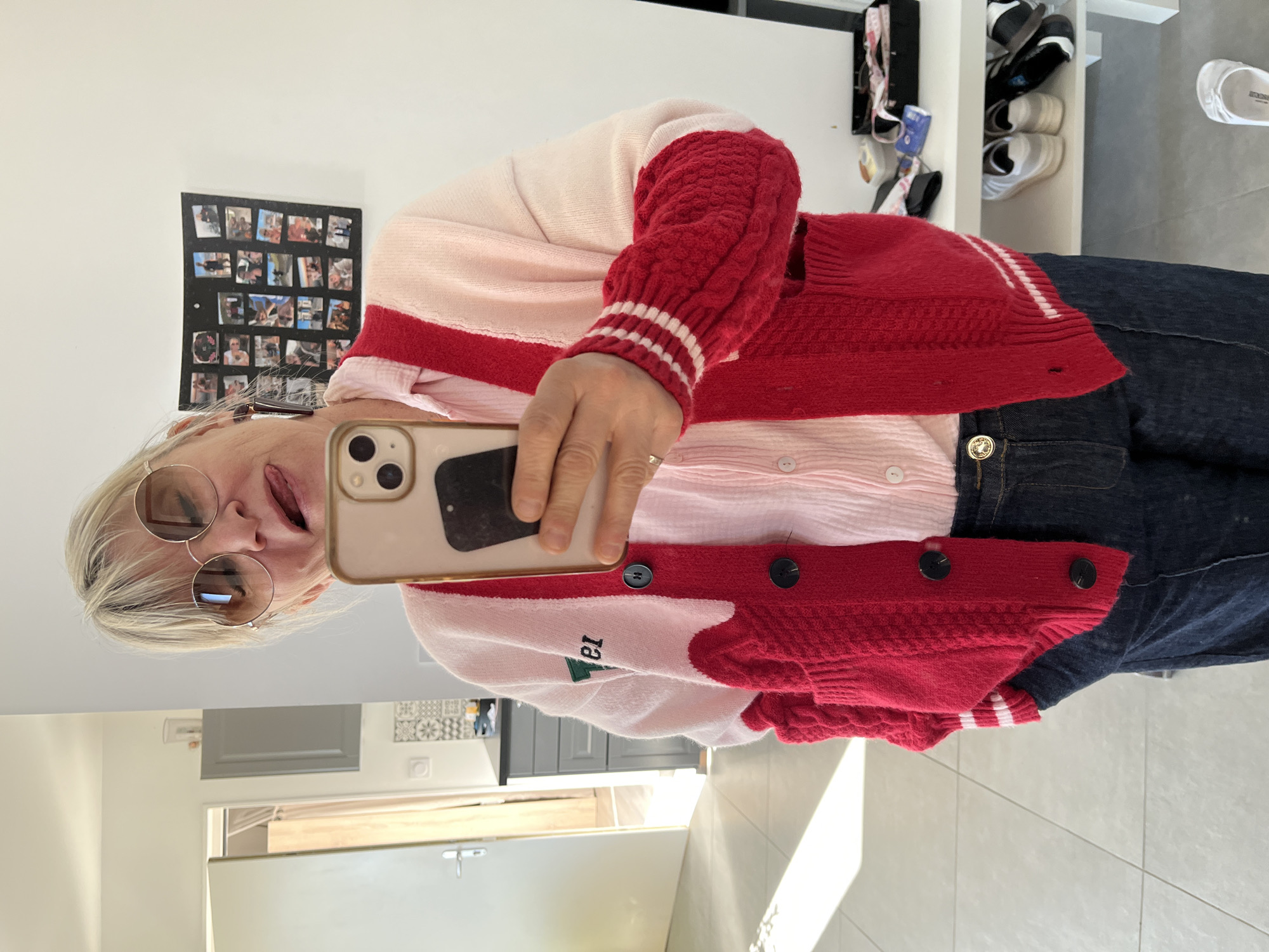 A blonde woman wearing round-frame glasses is taking a selfie in a mirror. She is wearing a pink blouse and a red and pink cardigan with the letters "13" on it. She also wears dark denim pants. The background shows a modern interior with a white wall adorned with a collage of photos and a white cabinet with shoes. The setting appears to be a home in Chambœuf, France.