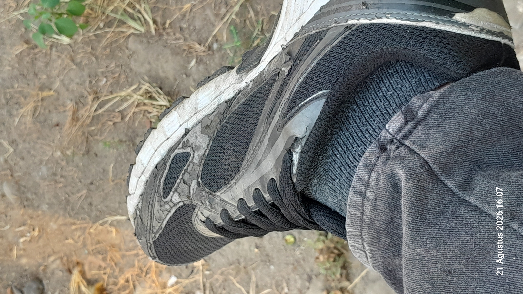 A worn, black and white athletic shoe is shown close-up, resting on gray pants. The shoe is dirty with soil and grass stains, and the sole shows signs of wear and tear. The background is blurred, consisting of dry grass and dirt. The bottom left corner of the image contains a timestamp that reads "21 Agustus 2026 16.07". This image was captured in Takeran, Indonesia.