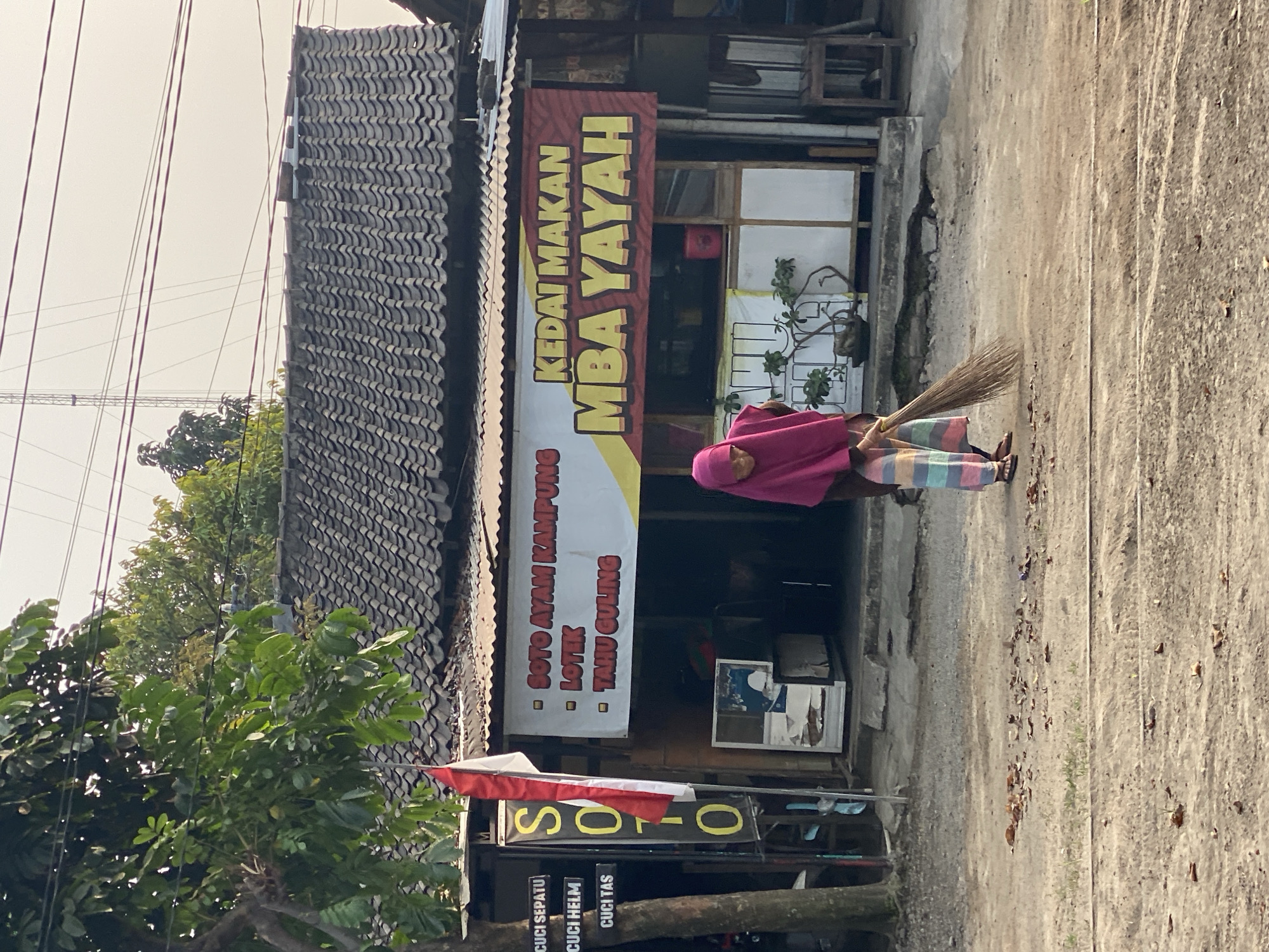 In the city of Kasihan, Indonesia, an elderly woman wearing a pink hijab and a striped skirt is sweeping the dusty ground in front of a small food stall. The stall, named "KEDAI MAKAN MBA YAYAH," displays a banner listing menu items such as "SOTO AYAM KAMPUNG," "LOTek," and "TAHU GULING." To the left of the stall, a vertical sign with the word "SOTO" is displayed alongside the Indonesian flag. The environment appears to be a street or pathway with a simple building with a tiled roof in the background, suggesting a local, casual dining establishment. The overall scene depicts a common, everyday activity in a commercial area.