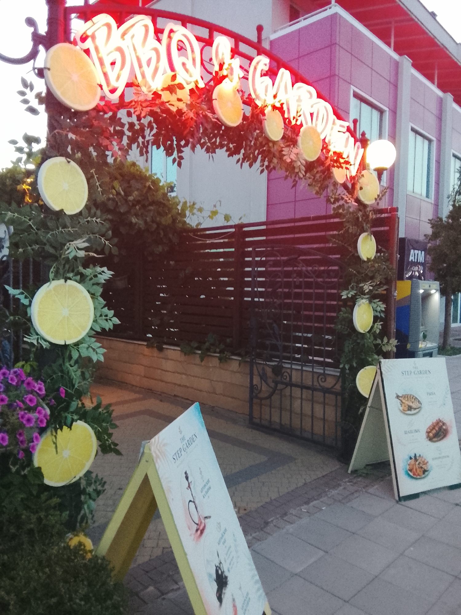 The image depicts an outdoor scene in front of a building in Nesebar, Bulgaria. An illuminated neon sign arches over an entrance, spelling out "BBQ & GARDEN" in red lettering. The sign is adorned with faux lemon slices and greenery. Below the archway, a dark metal fence encloses an area. To the right of the entrance, a standing sign displays menu items with images of food, including fish and grilled meats, and the text "THE STEP GARDEN." Another similar A-frame sign is partially visible to the left. A purple flower bush is situated near the left edge of the frame. An ATM is visible on the building in the background. The time of day appears to be late afternoon or early evening, as indicated by the lighting.
