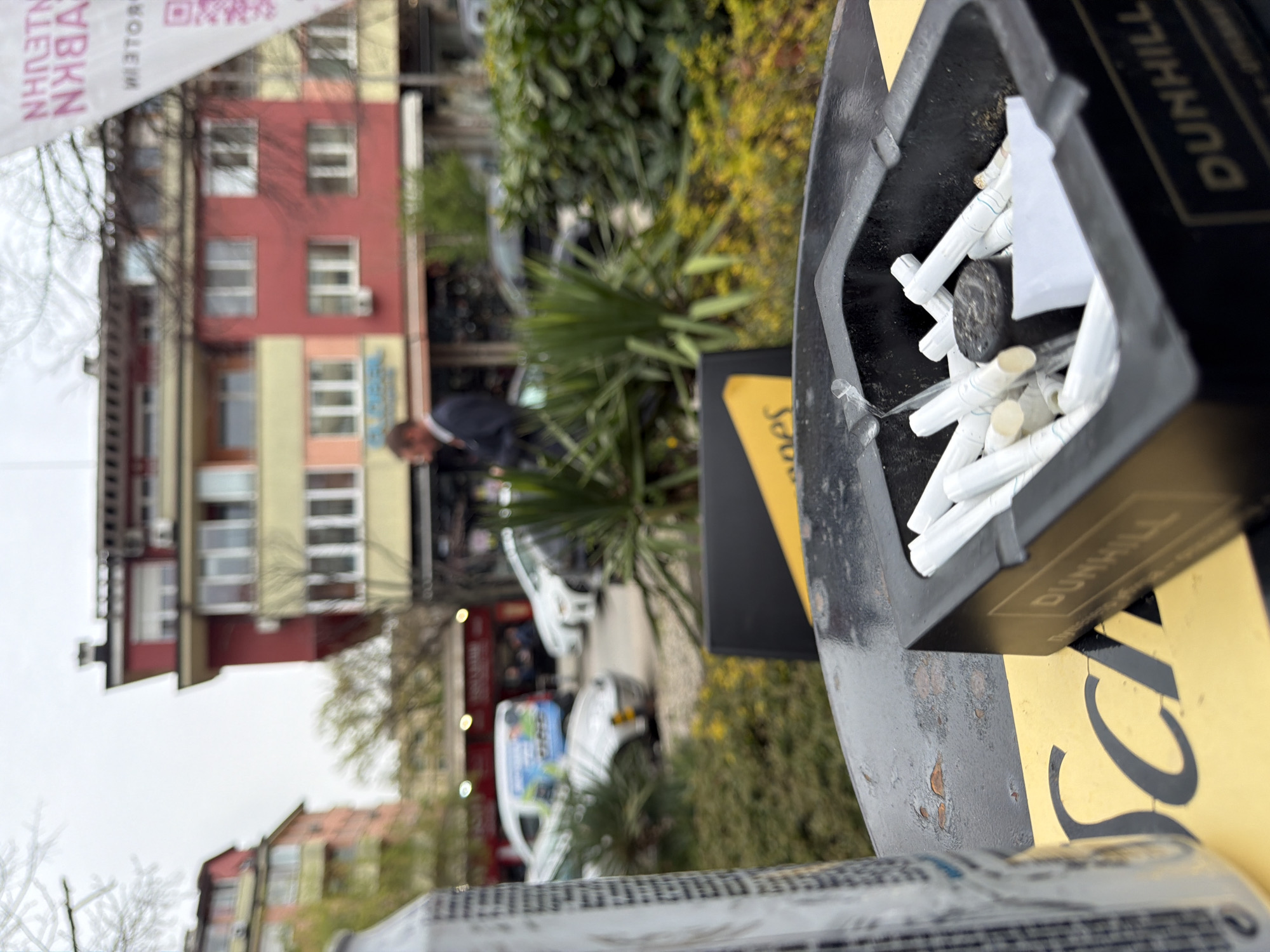 A close-up shot captures a cigarette disposal receptacle, seemingly made by the Dunhill brand, brimming with discarded cigarette butts. The bin is situated on a street in Sofia, Bulgaria. In the background, a blurred view reveals buildings and a street scene including a man in a suit and a van parked on the street. The image is taken at an angle and suggests an urban, outdoor setting. The bin's condition suggests wear and tear.