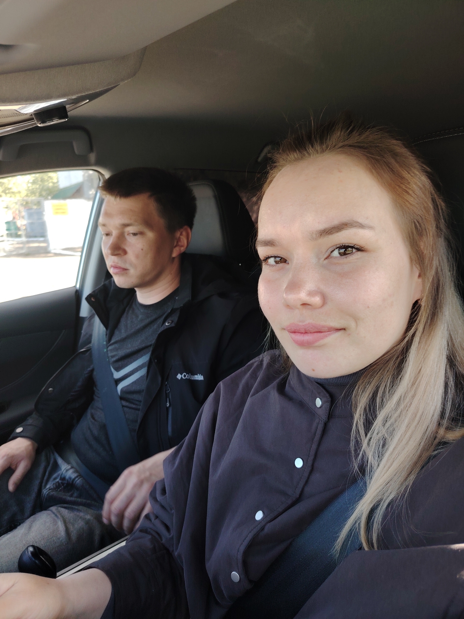 Two people are inside a car. The person in the foreground, a female with blonde hair, is wearing a dark jacket with buttons and a seatbelt. To her left is a male wearing a black Columbia jacket over a grey shirt and a seatbelt. Outside the car window, a street scene is visible with buildings and trees, suggesting the location is likely in a residential area. Given the prompt's specification, the car is within the city of Chaikovsky, Russia.