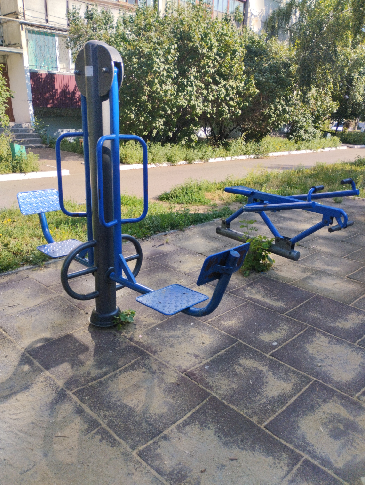 In the city of Kostanay, Kazakhstan, on a sunny day, outdoor exercise equipment is situated on a paved area. The equipment is primarily blue and gray, with a central gray post supporting several blue components. One section features two diamond-shaped blue footrests and handgrips, with a circular element at the base. Adjacent to this is another piece of equipment with two blue seats and a metal frame. The ground is a mix of paving stones and sand, with some patches of greenery. In the background, lush green trees and a multi-story building are visible. The scene appears to be a public park or recreational area.