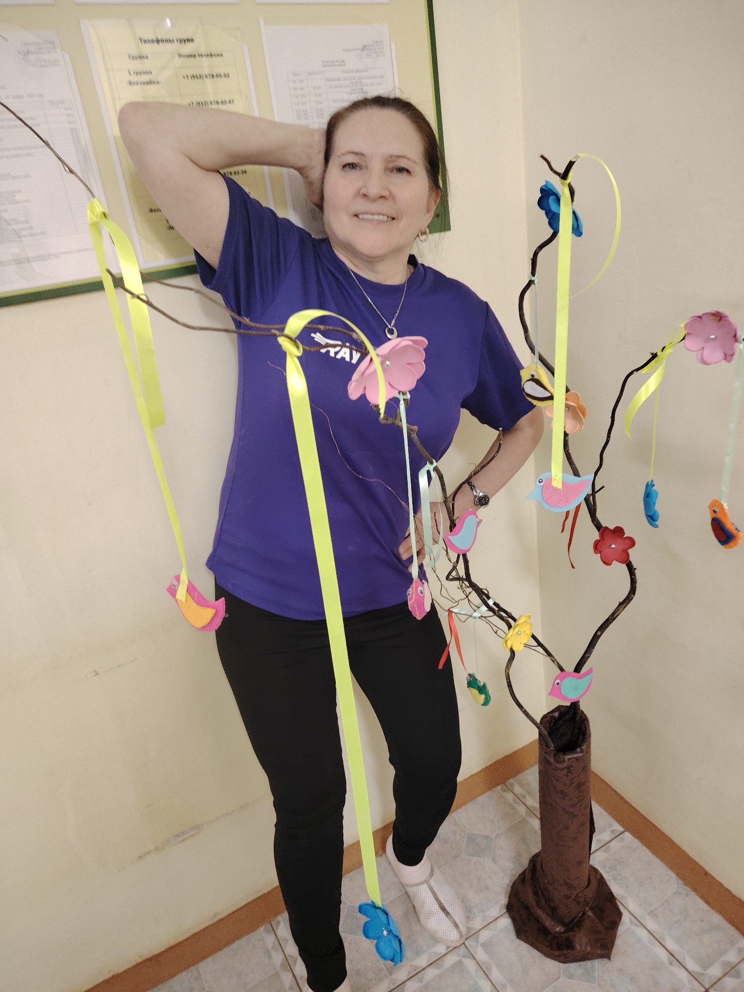 The image depicts a middle-aged woman standing indoors next to a decorative tree-like structure. She is wearing a purple t-shirt, black leggings, and white socks. Her left hand is resting on her head, while her right hand is on her hip. The tree-like structure is decorated with colorful paper flowers and bird-shaped ornaments hanging from yellow ribbons. A sign with telephone numbers is visible on the wall behind her. The location is suggested by the telephone numbers with country code +7, specifically identifying it in Russia, and based on the context, it's possibly in the city of Sarapul, Udmurtia Republic, where similar institutions can be found.
