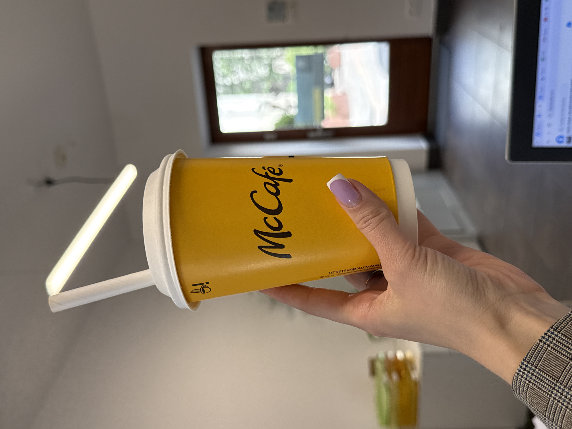 A hand with a french manicure holds a McCafe cup. The cup is yellow with black lettering. The text "McCafé" is visible in large, cursive font. Below this, smaller text reads "www.mcdonalds.pl" and some Polish characters that appear to be an address. A white paper straw is inserted into the cup, extending slightly beyond the rim. The background suggests an indoor setting with a window showing greenery and a desk with a monitor. A fluorescent light fixture is also visible. The image is likely taken during the daytime.