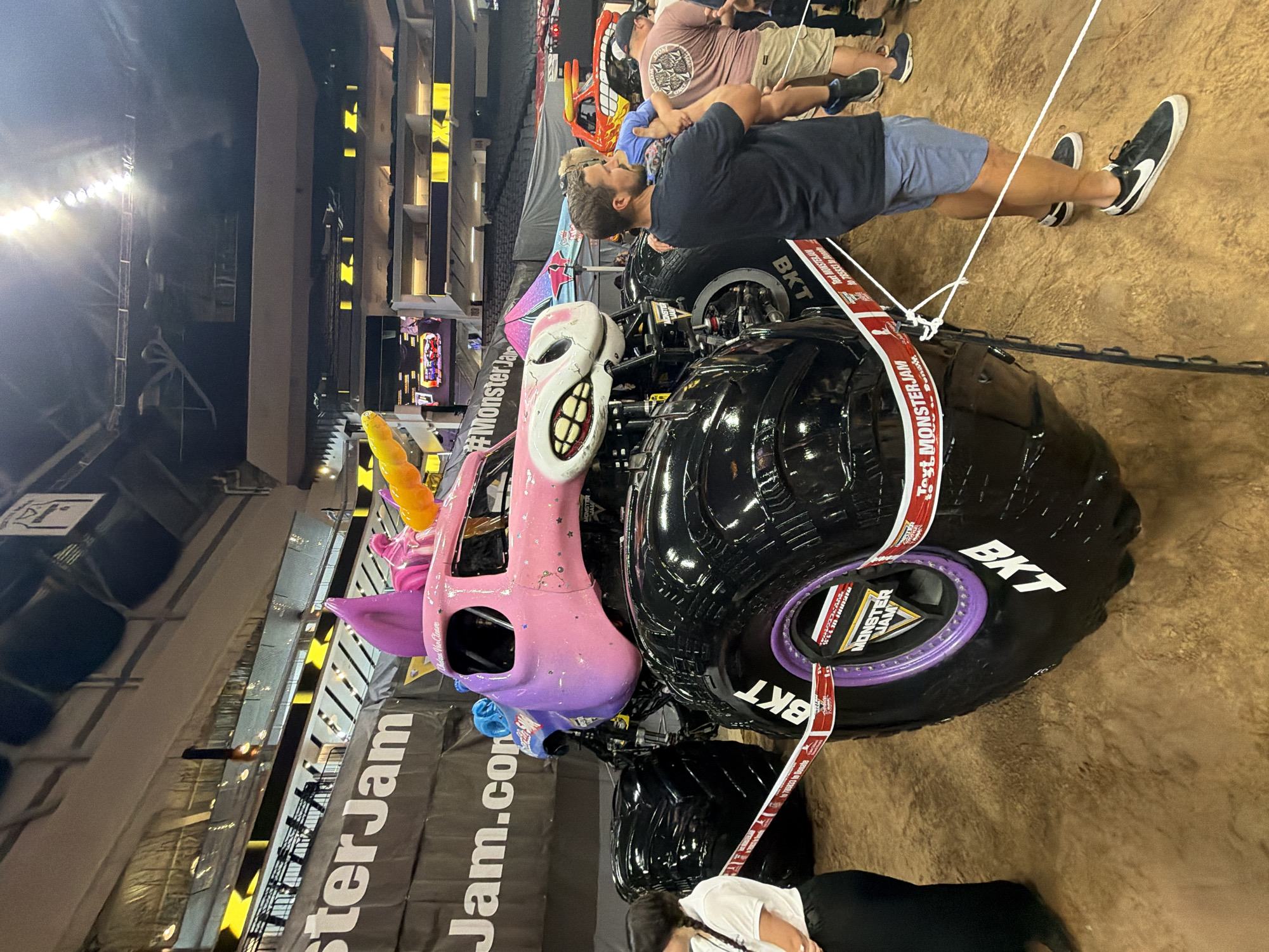 In Sacramento, California, an indoor arena hosts a Monster Jam event. The primary subject is a custom-built pink and purple monster truck designed to resemble a unicorn. The truck features large, black, deeply treaded tires with "BKT" emblazoned on them, and a purple rim. The front of the truck is sculpted into a unicorn's face with a large, open mouth revealing rows of white teeth and a pink tongue. A rainbow-colored horn is positioned on its head, and smaller purple ears are visible. The truck's body is adorned with glittery stars. A red and white ribbon with "Monster Jam" branding is draped across the front tire and around the truck, acting as a barrier. Several people are present. In the foreground, to the right, a man in a dark blue t-shirt and blue shorts holds a young child. Behind them, another person in a tan t-shirt and khaki shorts stands with a group of others. To the left, a person in a white shirt and black pants is partially visible. In the background, bleachers filled with spectators can be seen, along with overhead arena lighting and large banners with "Monster Jam" branding. The ground appears to be dirt or sand, typical of an arena floor for such events.
