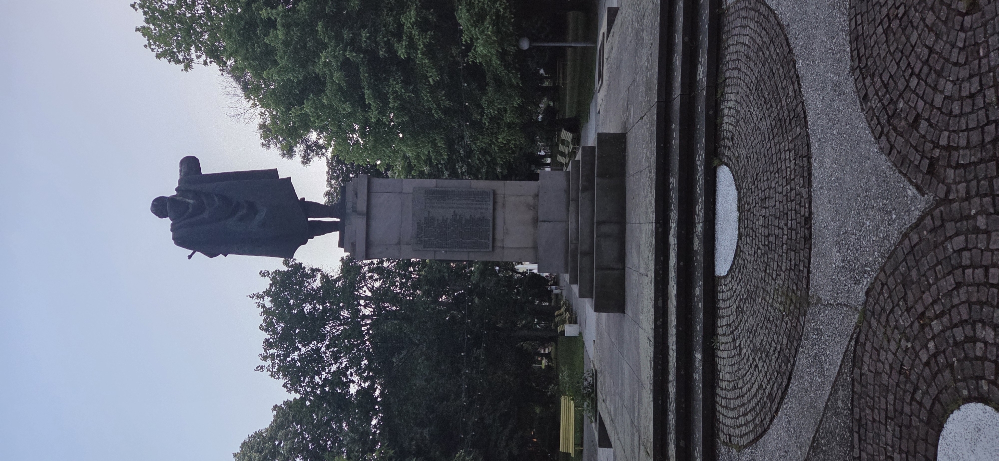 The image shows a statue of a man wearing a cloak, standing on a pedestal in an outdoor setting. The statue faces away from the camera, with its right arm extended. The pedestal is made of stone and has a plaque with text inscribed on it. In front of the pedestal are several stone steps leading down to a paved area with a circular pattern of cobblestones.
Surrounding the statue and pedestal are several large trees with lush green foliage. In the background, there are more trees, park benches, and lampposts. The sky is overcast and appears to be twilight or early morning.
This scene is located in the city of Parvomay, Bulgaria. The statue is likely a monument or memorial within a public park. There are no visible people in the immediate vicinity of the statue, though faint figures can be seen in the distance walking in the park. The overall impression is one of a somber and quiet public space.