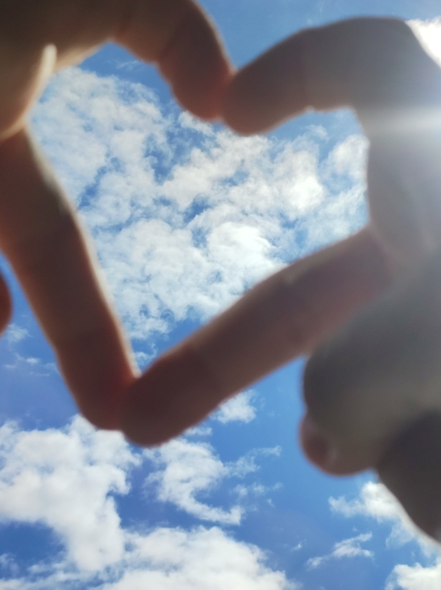 The image shows a pair of hands forming a heart shape against a backdrop of blue sky with scattered white clouds. The hands are out of focus. A bright light source, possibly the sun, is visible in the upper right corner. There is no indication of the location being in Chaikovsky, Russia, or any other specific location.