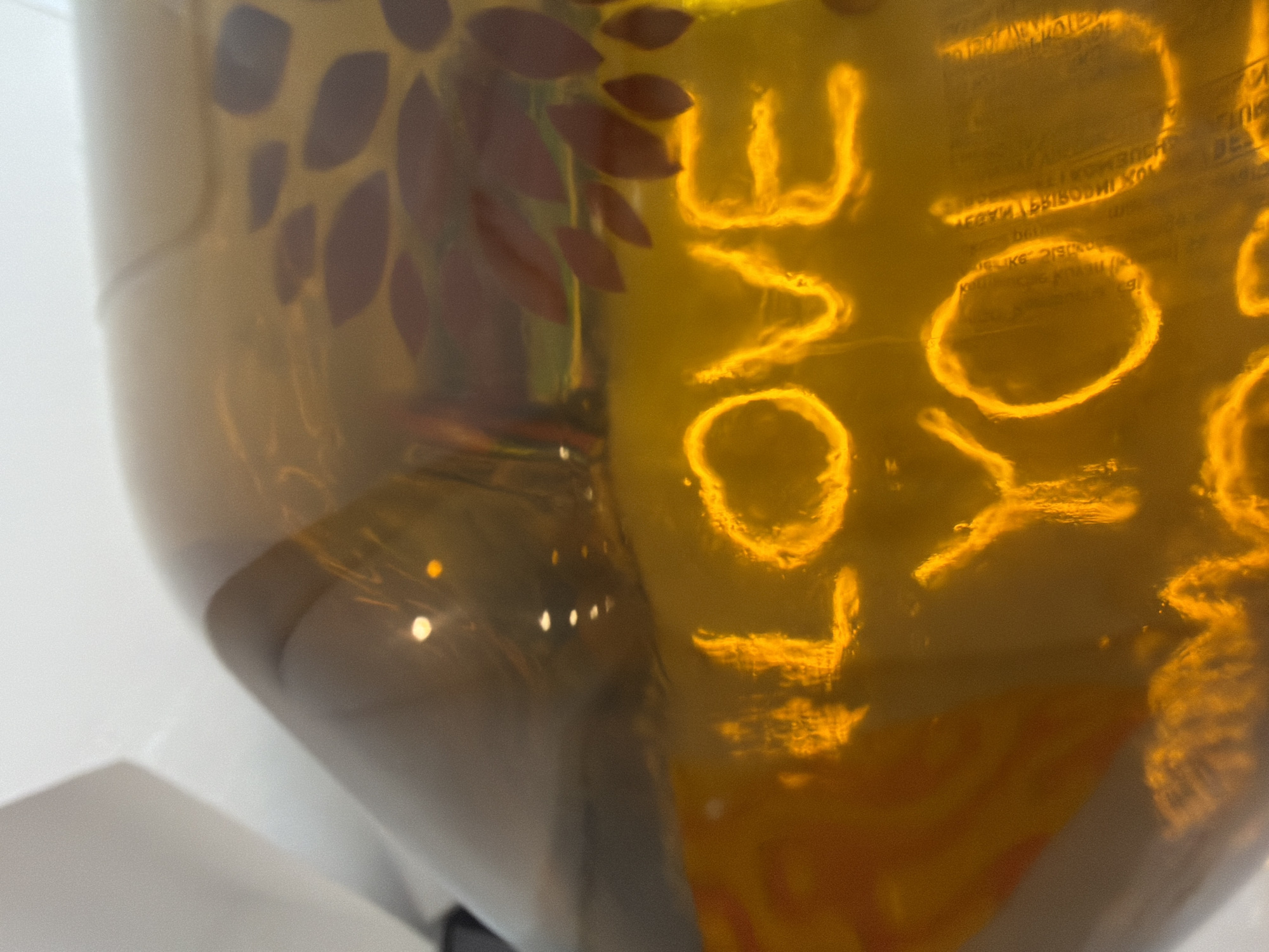 The image shows a close-up of a translucent, possibly plastic, container with lettering on its surface. The lettering appears to say "LOVE YOU", in vertically stacked words. The letters are in a bright, possibly neon, yellow-orange color.
On the left side of the container, there are blurred images of what seems to be leaves or petal-shaped designs, darker in color, seemingly attached or printed on the container's surface.
The background appears to be a simple white surface. The reflection of light suggests the container is smooth and glossy.
Without further context, it is impossible to determine the location in Budva, Montenegro, or identify any people, actions, or interactions related to the image.