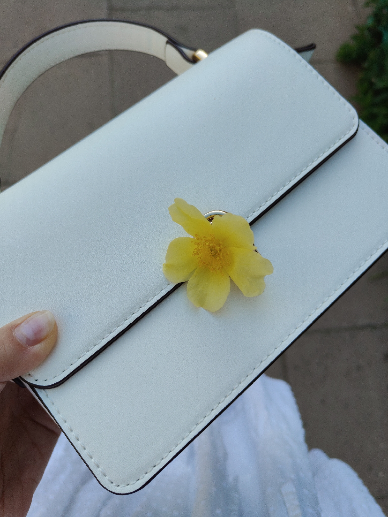 The image shows a person holding a white handbag with a single yellow flower placed on top of the clasp. The handbag has a structured shape with a handle and a dark trim. The person's hand is visible in the lower-left corner, with a glimpse of white fabric near the bottom of the frame. The background appears to be an outdoor setting with some pavement visible, and greenery in the upper-right corner. The location of Unknown, Unknown, is not discernible from the image.