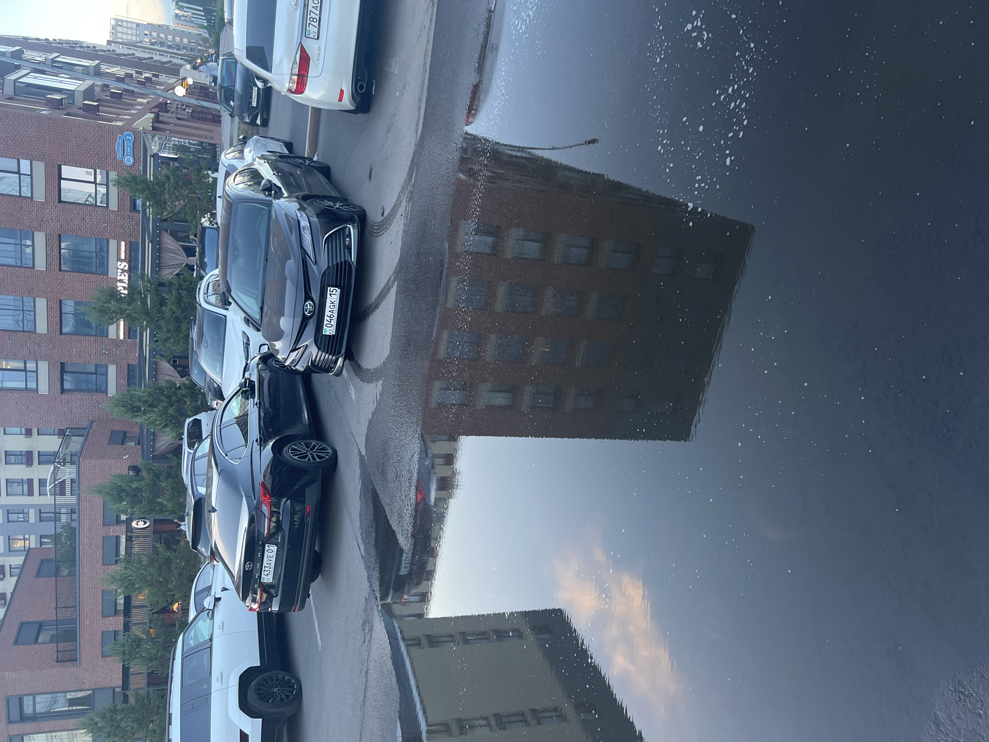 A paved parking lot at dusk in Astana, Kazakhstan features several cars, including a white Range Rover, a black Lexus sedan, and a dark gray Toyota Camry. The vehicles are parked in front of a multi-story brick building with numerous windows. A large puddle of water reflects the building and the cloudy sky. A sign with "PEOPLE'S" written on it is visible on the building. The ground is wet, indicating recent rain or cleaning. The scene is calm and devoid of any visible activity.