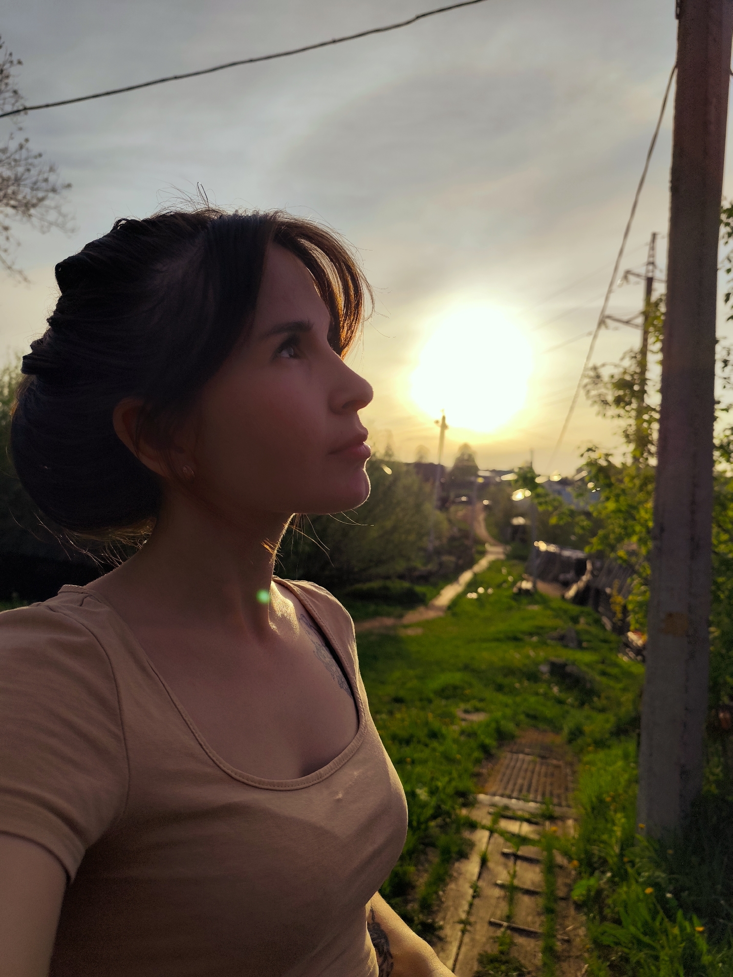 A medium shot captures a woman looking upwards towards the setting sun. Her brown hair is styled up with bangs framing her face. She is wearing a light brown t-shirt, and a tattoo is visible on her upper arm. The background reveals a rural landscape with green grass and a path made of wooden planks leading through it. In the distance, houses and utility poles are silhouetted against the bright sun. The location is likely Фоки, Russia.