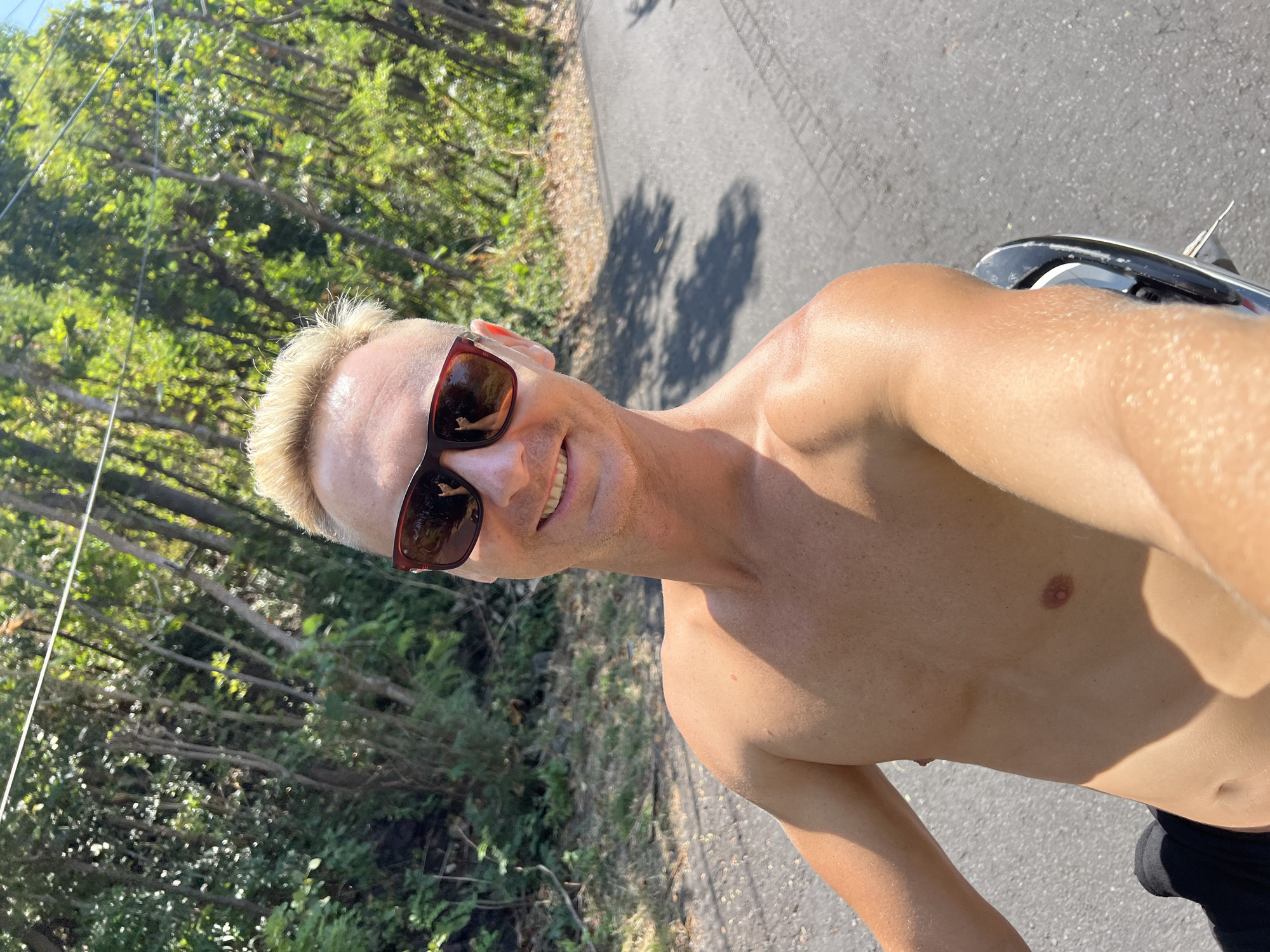 A shirtless man with blonde hair and sunglasses is taking a selfie while on a scooter. He is smiling and looking at the camera. The background is a road lined with trees. The scene appears to be in Abang, Indonesia, given the lush tropical vegetation. The lighting suggests it is daytime.