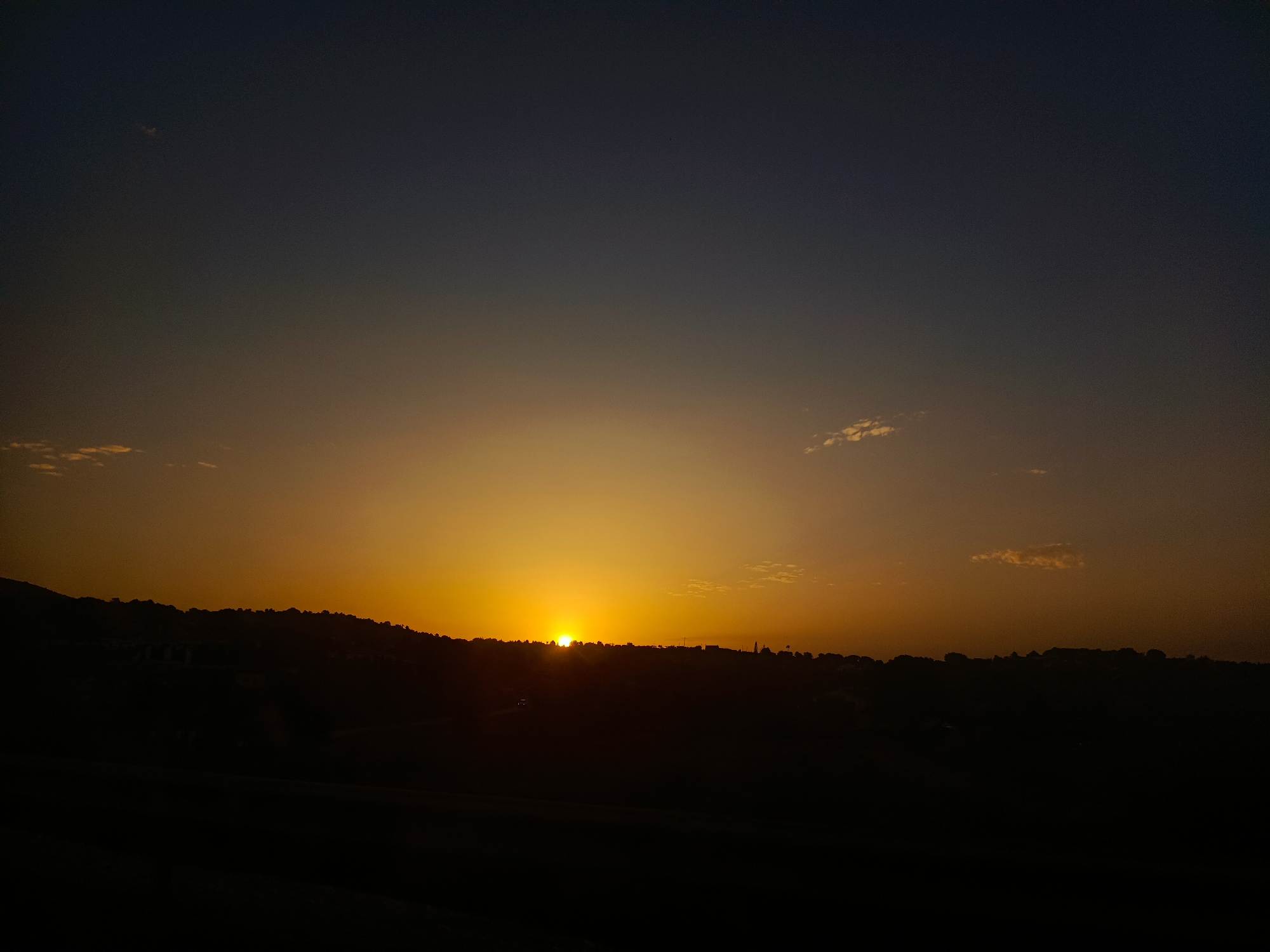A wide shot captures the dramatic silhouette of a sunset over the town of Senija, Spain. The sky transitions from a deep indigo at the top to a vibrant golden orange near the horizon. A sliver of the sun peeks over the dark, undulating line of hills that form the town's backdrop. Tiny dots, likely distant buildings or trees, are visible along the crest of the hills. The foreground is a solid expanse of black, obscuring any details of the ground or immediate surroundings. A few wispy clouds, tinged with orange, are scattered in the upper sky. The overall impression is one of natural beauty and the quiet descent of day in a Spanish town.