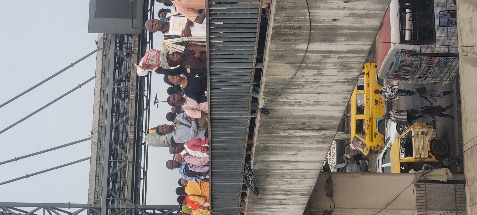 The image shows a street scene in Lagos, Nigeria, viewed from an elevated position, possibly a pedestrian bridge or building. A crowd of people are standing on the bridge, looking towards something out of frame. Below the bridge, there is traffic, including several yellow and white buses typical of Lagos. There are people walking on the street adjacent to the vehicles. Advertisements are visible on the sides of the buses and buildings. The sky is overcast.