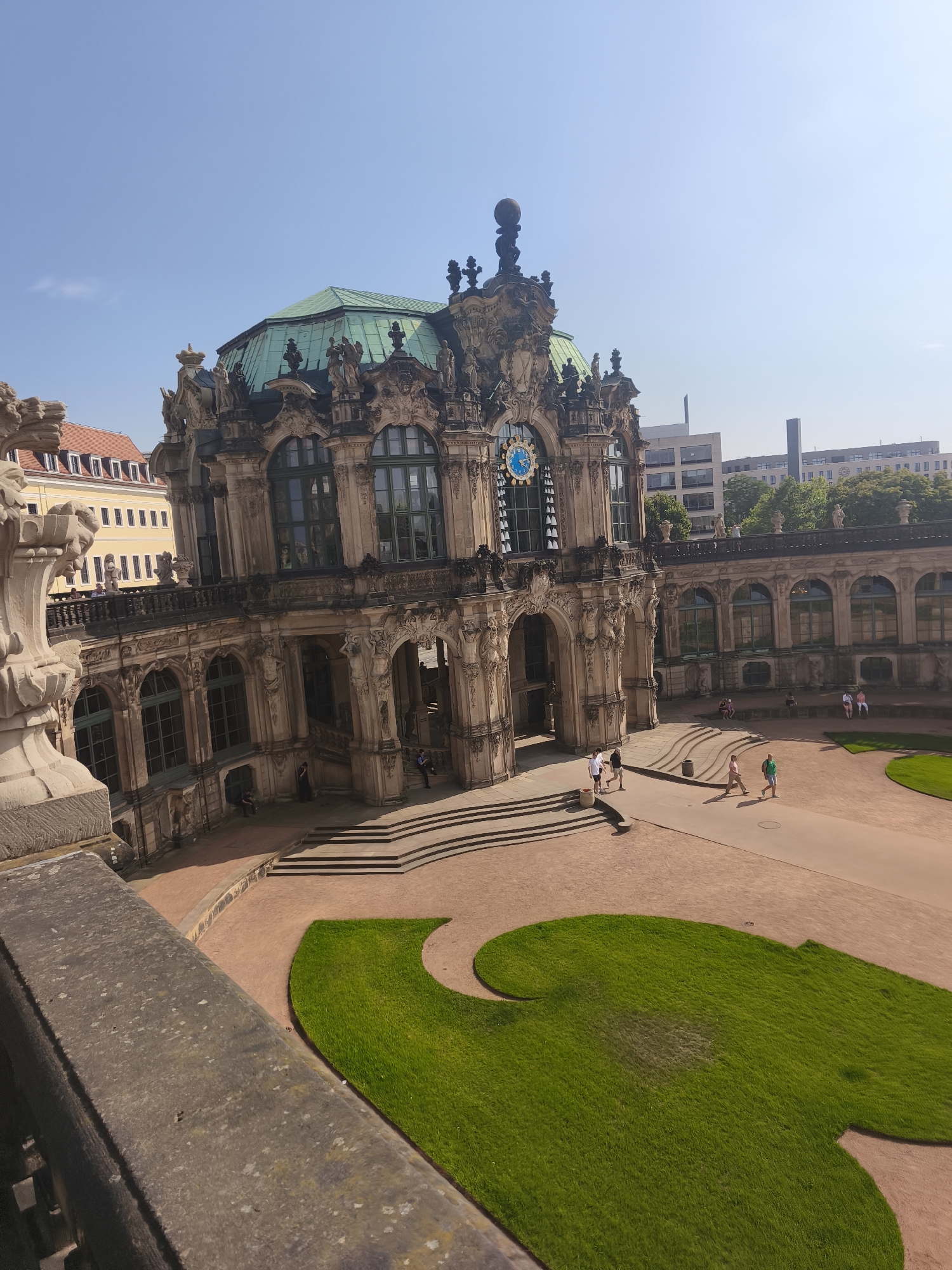 The image shows a high-angle view of the Zwinger, a Baroque palace in Dresden, Germany. The main building, with its ornate facade and green dome, is prominently featured. A clock with a blue face is visible on the building's central tower. In the courtyard, several people are walking and sitting. A stylized green lawn is situated in the foreground. The sky is clear and blue.