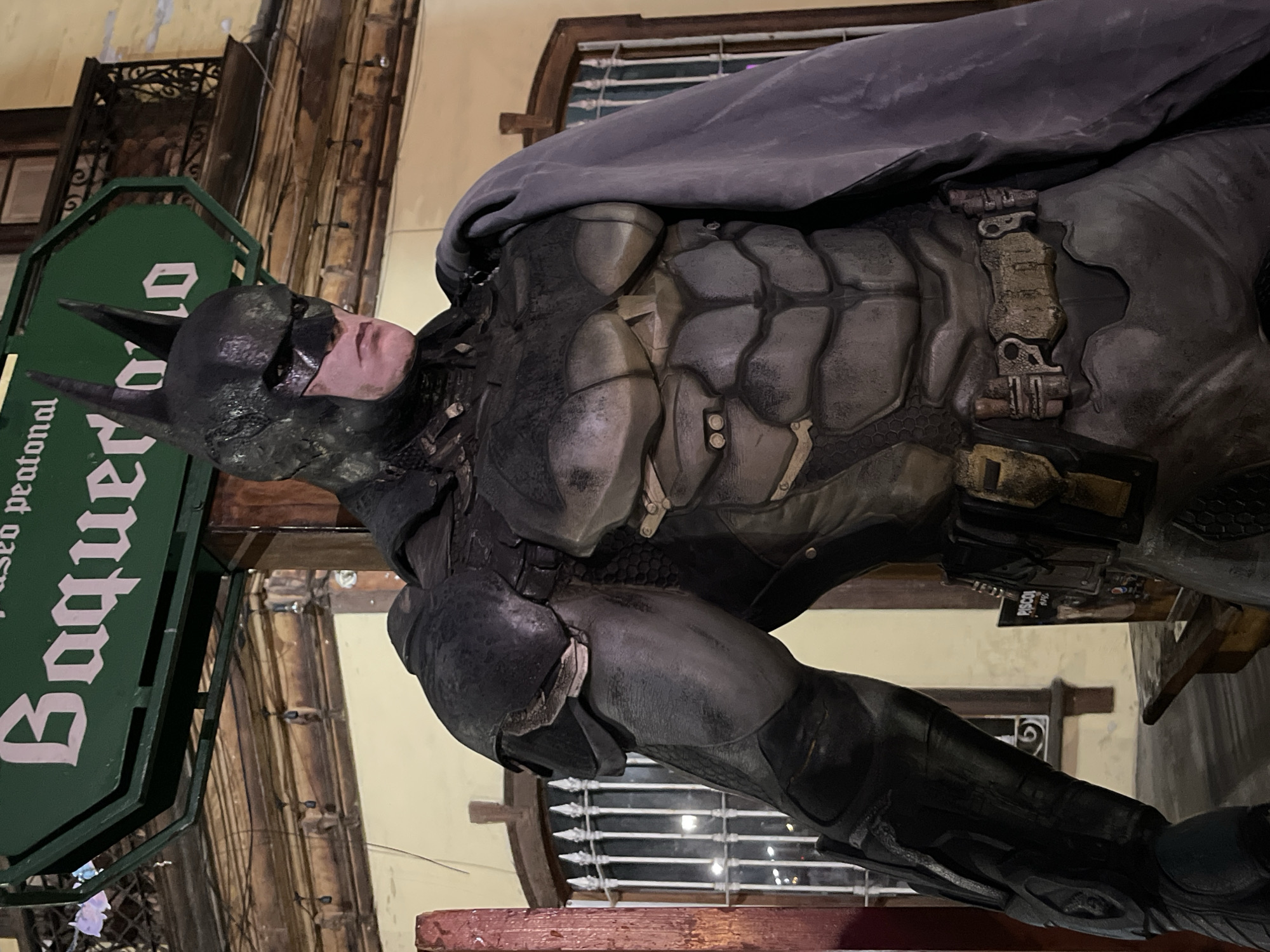 A statue of Batman is standing on a red-painted surface in front of a building. The statue is clad in a heavily textured, armored suit with a dark gray cape. The mask and cowl are black, and part of a face is visible, though it appears to be made of plastic or a similar material. To the left of the statue is a green sign with white text that reads "Baqueta paseo peatonal" vertically. The building behind the statue features architectural details like columns and windows with horizontal bars. The location seems to be an urban area, based on the architectural style of the buildings.