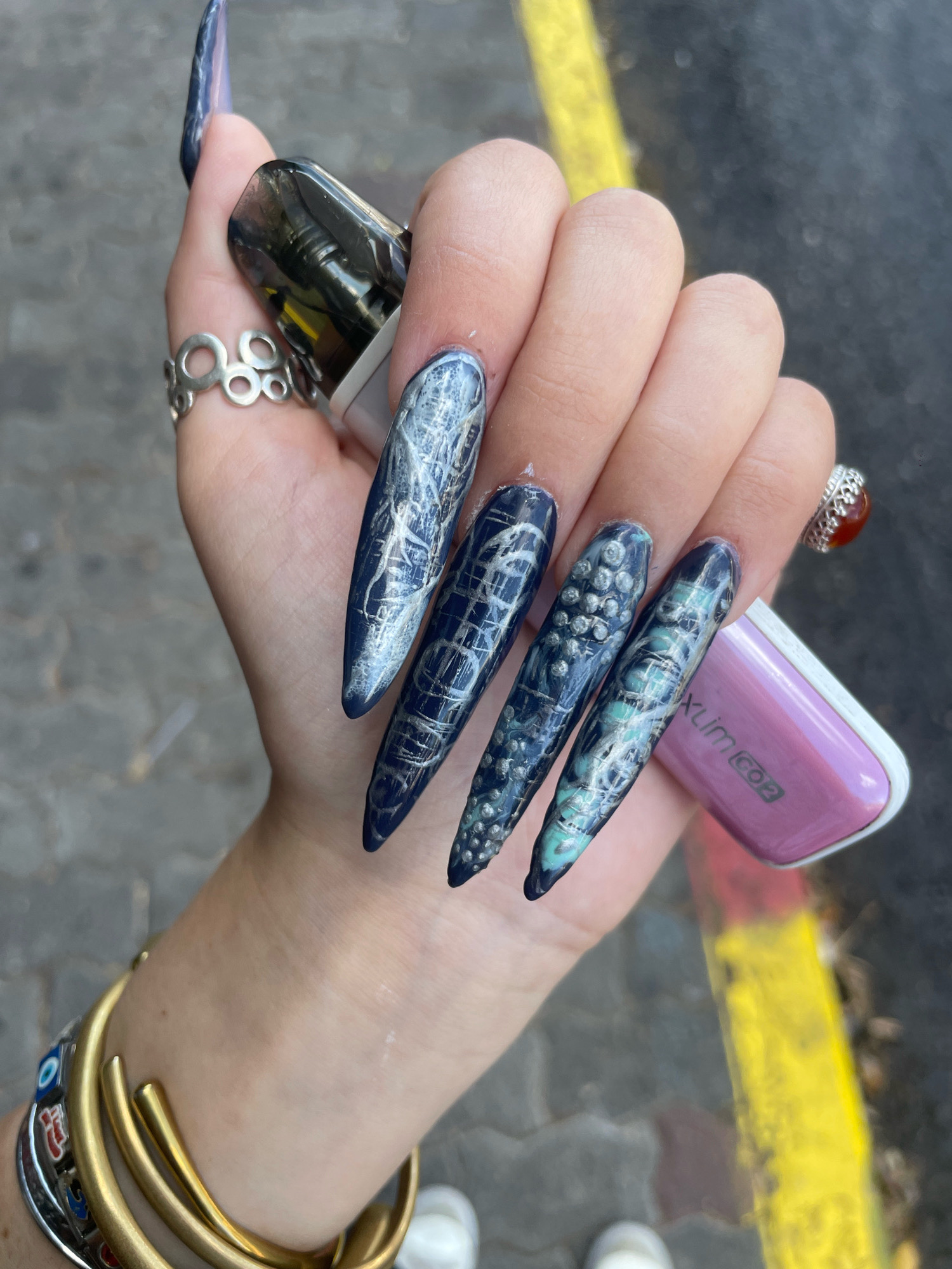 A close-up, eye-level shot shows a person's left hand, adorned with elaborate stiletto-shaped fingernails featuring a dark blue base with intricate silver and blue-green designs. The fingernails appear to have a textured, almost scaly pattern on some, and a cracked or web-like pattern on others. The hand is holding a dark-colored vaping device in the thumb and forefinger, and a small, light purple vaping device with the text "Xlim C02" is visible near the pinky finger. The person is wearing several rings on their fingers, including a silver ring with interconnected circles on the index finger and a silver ring with a red stone on the ring finger. On the wrist, a thick gold cuff bracelet and a layered bracelet with multiple charms, including a blue evil eye, are visible. The background consists of a textured gray surface, likely pavement, with a bright yellow line running horizontally across the lower right portion of the frame, suggesting an outdoor urban environment. The scene is consistent with an outdoor setting in a city, possibly Raanana, Israel, given the visible yellow line marking, which is common in Israeli urban planning.