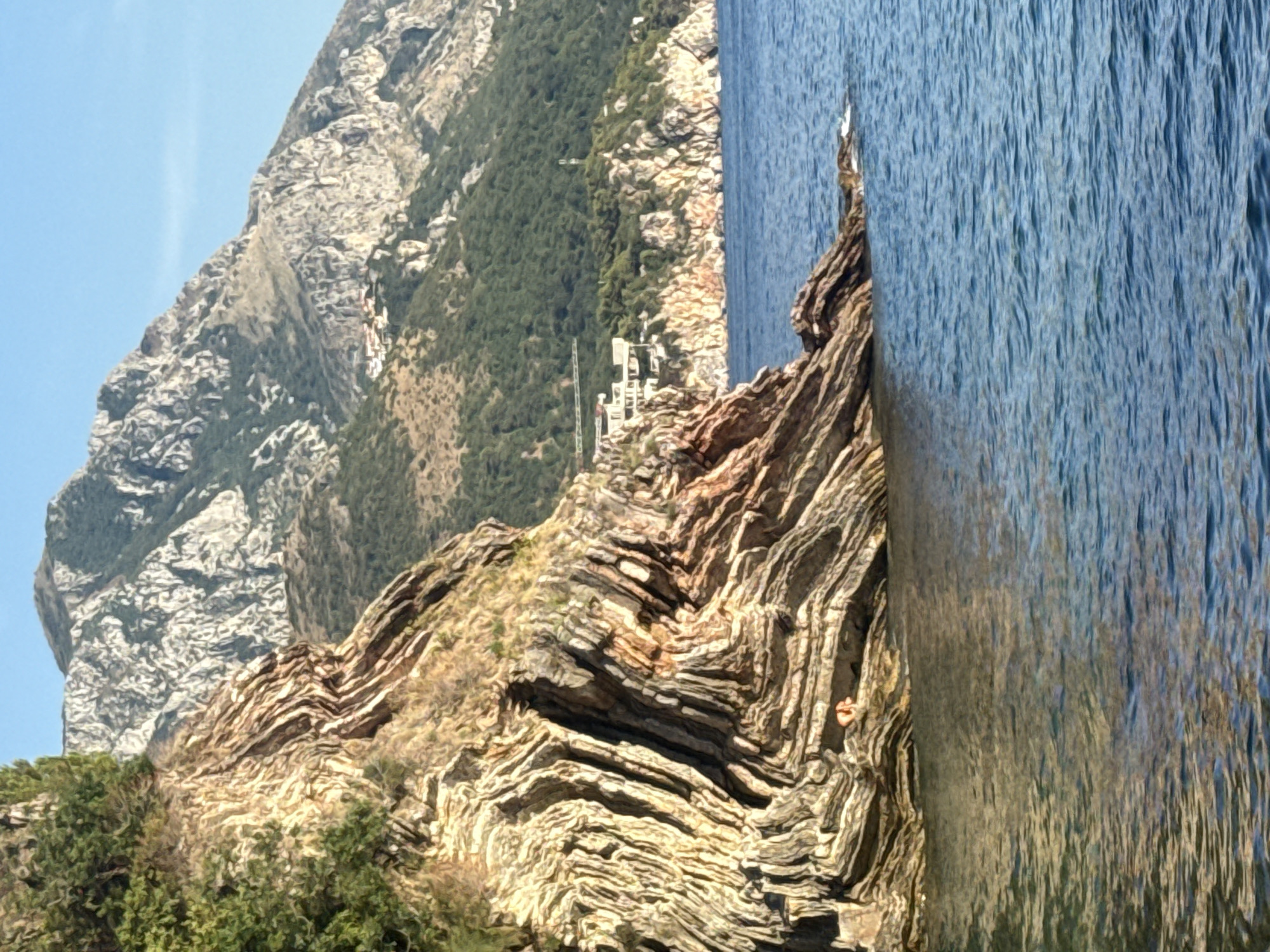 This image depicts a coastal scene in Rafailovići, Montenegro, characterized by dramatic geological formations and the Adriatic Sea. In the foreground, the sea displays a deep blue hue with gentle ripples reflecting the sunlight. The focal point is a rugged, layered cliff face composed of sedimentary rock, exhibiting striking striations and folds in shades of tan, brown, and reddish-orange. Lush green vegetation clings to the upper portions of this cliff.
In the middle ground, the cliff gives way to the sea, with a rocky outcrop jutting into the water. Further in the distance, a densely forested hillside slopes upwards towards formidable, rocky mountains that dominate the background. A cluster of white buildings, possibly residential or tourist accommodations, is visible nestled on the hillside among the trees. The sky above is a clear, bright blue, suggesting a sunny day. A single person, appearing as a small, orange-clad figure, is seated on a ledge of the cliff face, near the waterline.