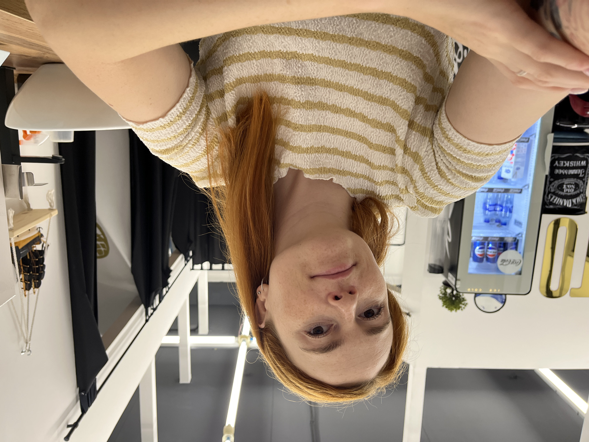 A woman with shoulder-length, reddish-blonde hair and fair skin with freckles is looking directly at the camera. She is wearing a white and gold horizontally striped top with a textured, smocked pattern. Her hands are clasped in front of her. In the background, to her left, is a Coca-Cola branded refrigerator stocked with drinks. To her right, a white ceramic bowl sits on a wooden surface. Black curtains hang in the background. The environment appears to be an indoor commercial space, possibly a shop or salon. The location is not Chaikovsky, Russia.