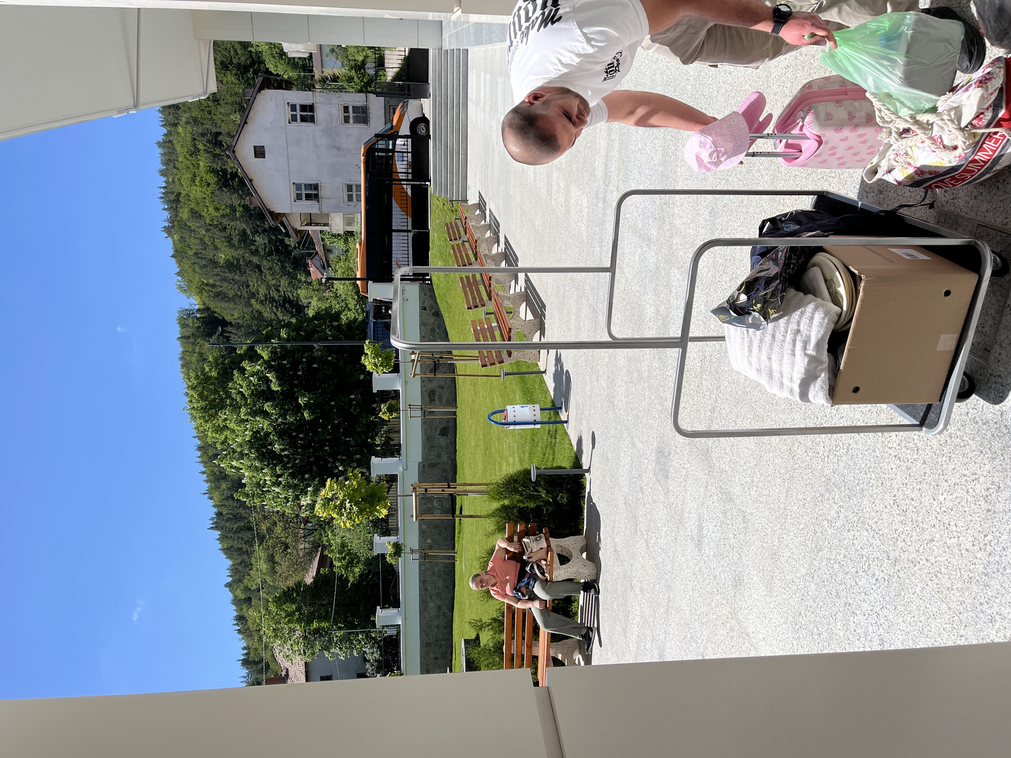 A person wearing a white t-shirt and khaki pants is adjusting a pink hat on top of a pink suitcase that is sitting on a luggage cart, which is also loaded with other luggage, on a light-colored stone-paved area. In the background, there are benches arranged on a grassy area, and a person is sitting on one of the benches. Behind the benches, there is a building and a forested area. The scene appears to be outside a building. The location is Belchinski Bani, Bulgaria.