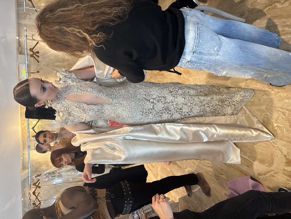 This image, rotated 90 degrees clockwise, captures a bustling behind-the-scenes moment, likely in a dressing room or studio, where models are being prepared.
The central figure is a female model with light skin and dark hair styled in a sleek, pulled-back bun. She wears an exquisite, floor-length gown made of intricate cream or light beige lace, featuring a high neck and decorative ruffled details on the shoulders. The dress has striking cutouts on the sides of the torso, adding a contemporary touch. She is adorned with large, ornate, gold-toned statement earrings. Her gaze is directed slightly to her left, with a neutral expression.
Standing directly behind and to the right of the main model, with her back to the viewer, is a person with long, voluminous, curly brown hair. She is dressed in a black long-sleeved top and light blue, distressed jeans with paint splatters. She appears to be adjusting the main model's dress or handling the voluminous, shimmering cream-colored satin fabric that is draped around the model's lower body and extending onto the floor.
Further back in the scene, another female model with dark hair is visible, wearing a partially seen white or sheer garment, possibly also a lace dress. Her head is tilted slightly, and her gaze is directed towards the camera. Behind her, the head of another individual with dark hair is partially visible.
To the left of the main model, closer to the foreground, a person with reddish-brown hair is seen kneeling or sitting, wearing a dark top. In the bottom left corner, a person with a dark skin tone is partially visible, wearing a light-colored top with straps and studded detailing, paired with dark pants and a wide, studded belt. On the bottom right, another individual's hands and forearms are visible, suggesting they are also working or present in the space. Several plastic water bottles are visible on the floor nearby.
The setting appears to be indoors, likely a professional preparation area given the array of models and stylists. The floor is made of large, polished marble tiles with light beige and brown veining. In the background, a clothing rack with various garments is partially visible, further suggesting a fashion-related context. The lighting is bright and even, illuminating the scene clearly. The overall impression is one of active preparation for a fashion show, photoshoot, or formal event. No discernible text is visible in the image.