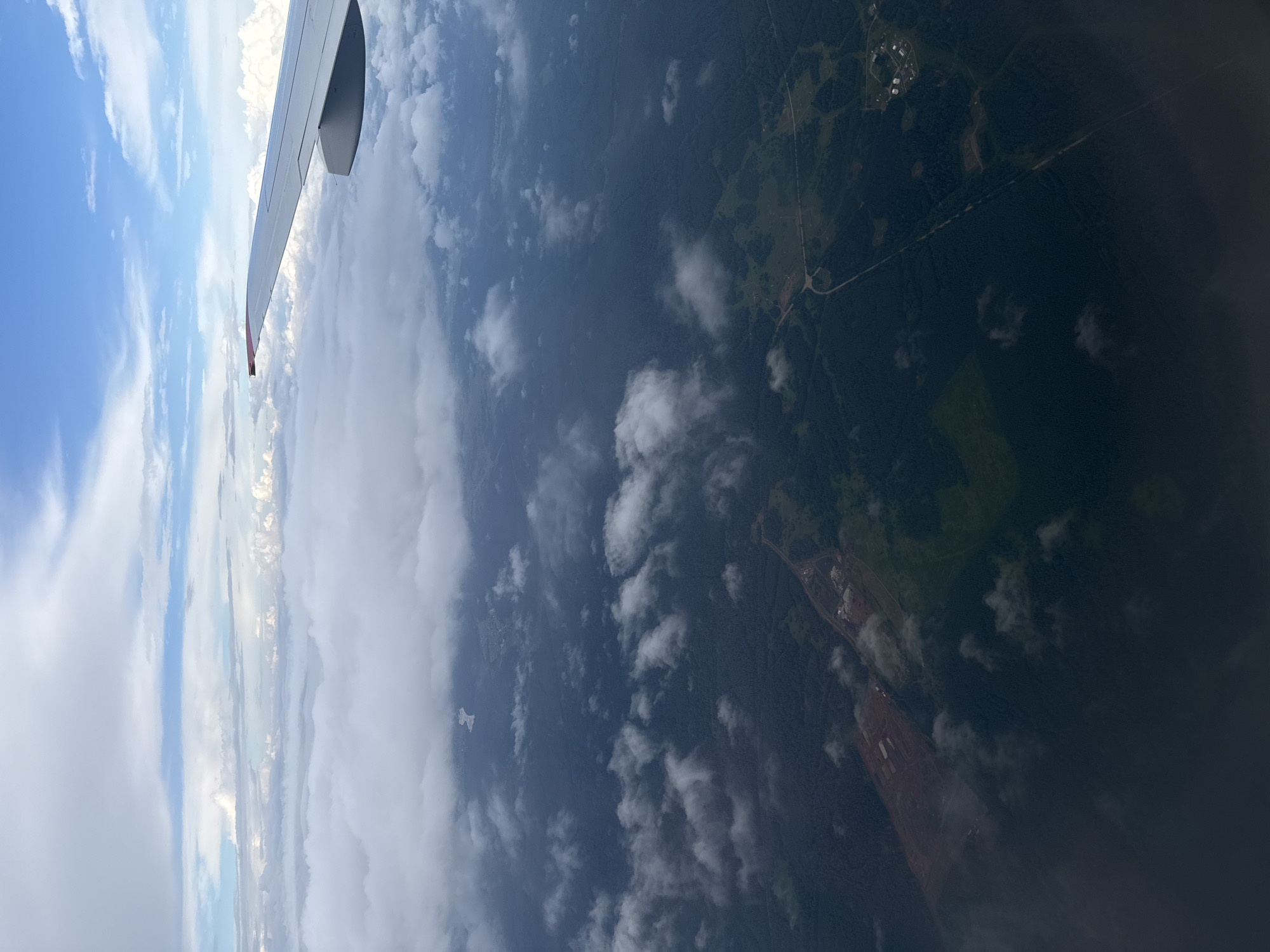 The image is an aerial view of a landscape, likely captured from an airplane. In the upper right quadrant, a portion of an airplane wing is visible. The sky is a mix of blue and white clouds, with a thin layer of clouds obscuring the ground in places. The landscape below appears to be predominantly forested, with some areas of cleared land and a road visible. There is a small settlement or group of buildings visible in the upper right portion of the forest. The overall impression is one of a rural or remote area seen from a high altitude. There are no people or specific actions depicted. The lighting suggests it is daytime.