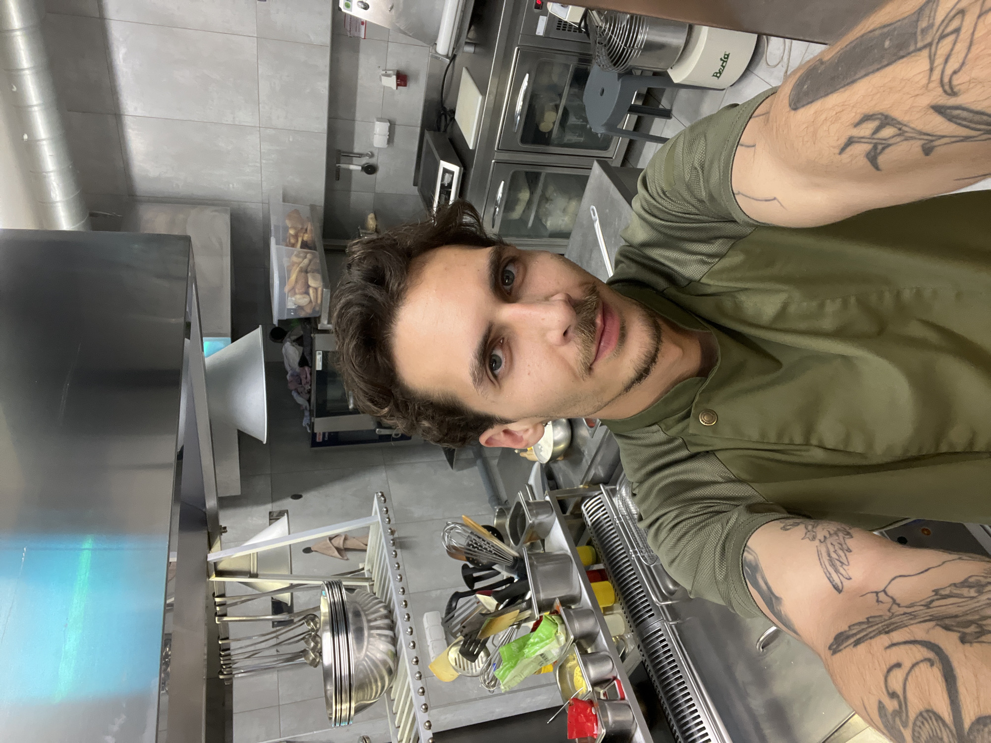 A man with brown hair, light eyes, and a mustache is taking a selfie in what appears to be a commercial kitchen. He is wearing a green chef's coat and has tattoos on his arms. Stainless steel equipment, including pots, pans, utensils, and ovens, is visible in the background. The location is potentially a kitchen in Budva, Montenegro.