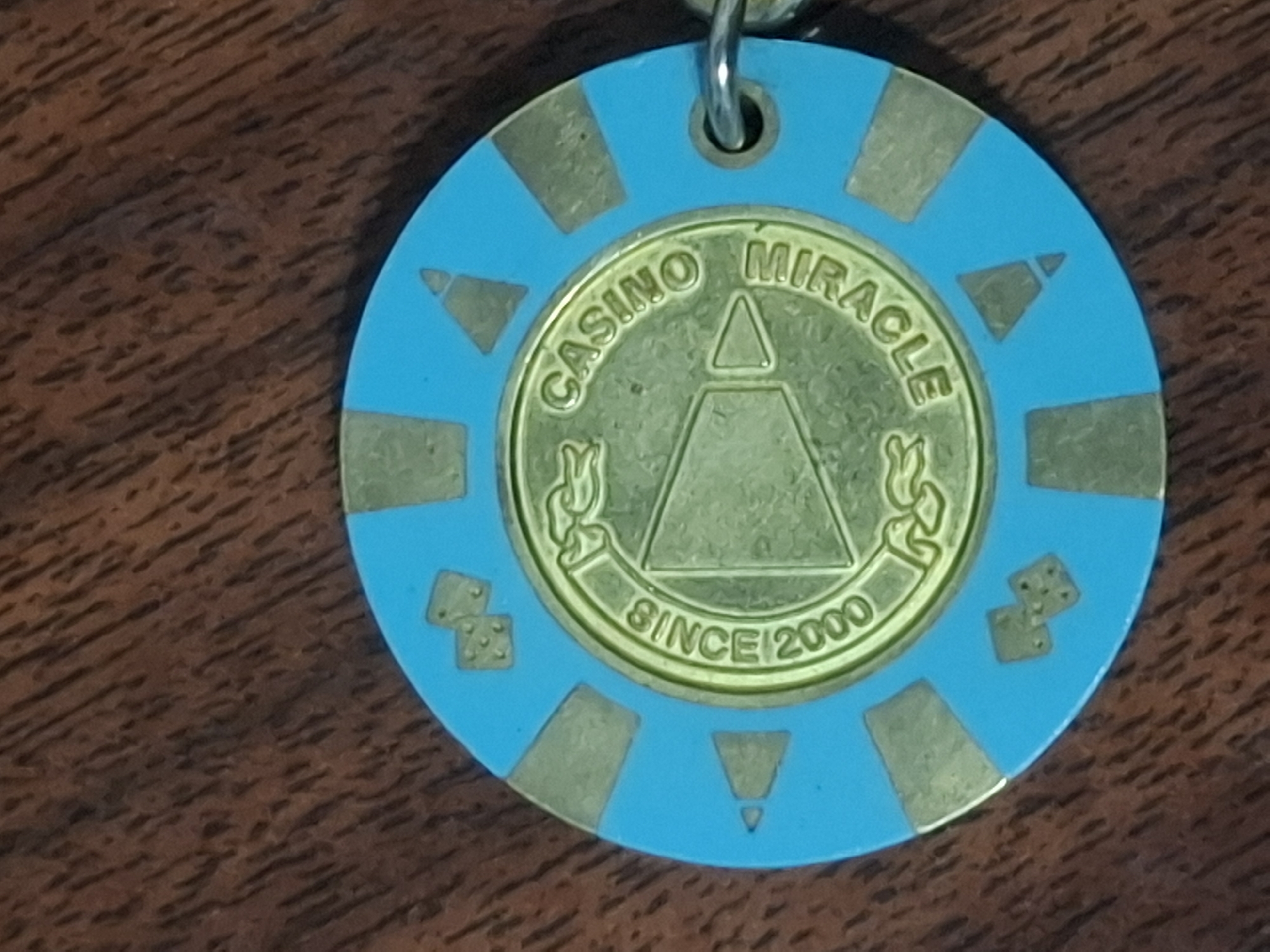 A blue and gold casino chip rests on a dark brown wooden surface. The chip features a circular gold inlay with embossed text and imagery. The text "CASINO MIRACLE" is printed in an arc around the top of the inlay, with "SINCE 2000" printed in an arc around the bottom. Inside this text, a graphic depicts a stylized megaphone or speaker. The outer edge of the chip is bright blue, punctuated by alternating gold rectangular inlays and gold triangular shapes. Two gold dice symbols are visible on the upper left and lower right portions of the blue outer rim. A small silver keychain ring is attached to the right side of the chip. The image is a close-up shot, with the chip dominating the frame. The background is a close-up of wood grain. No people or animals are present in the image. The location in Almaty, Kazakhstan is not ascertainable from the provided image.