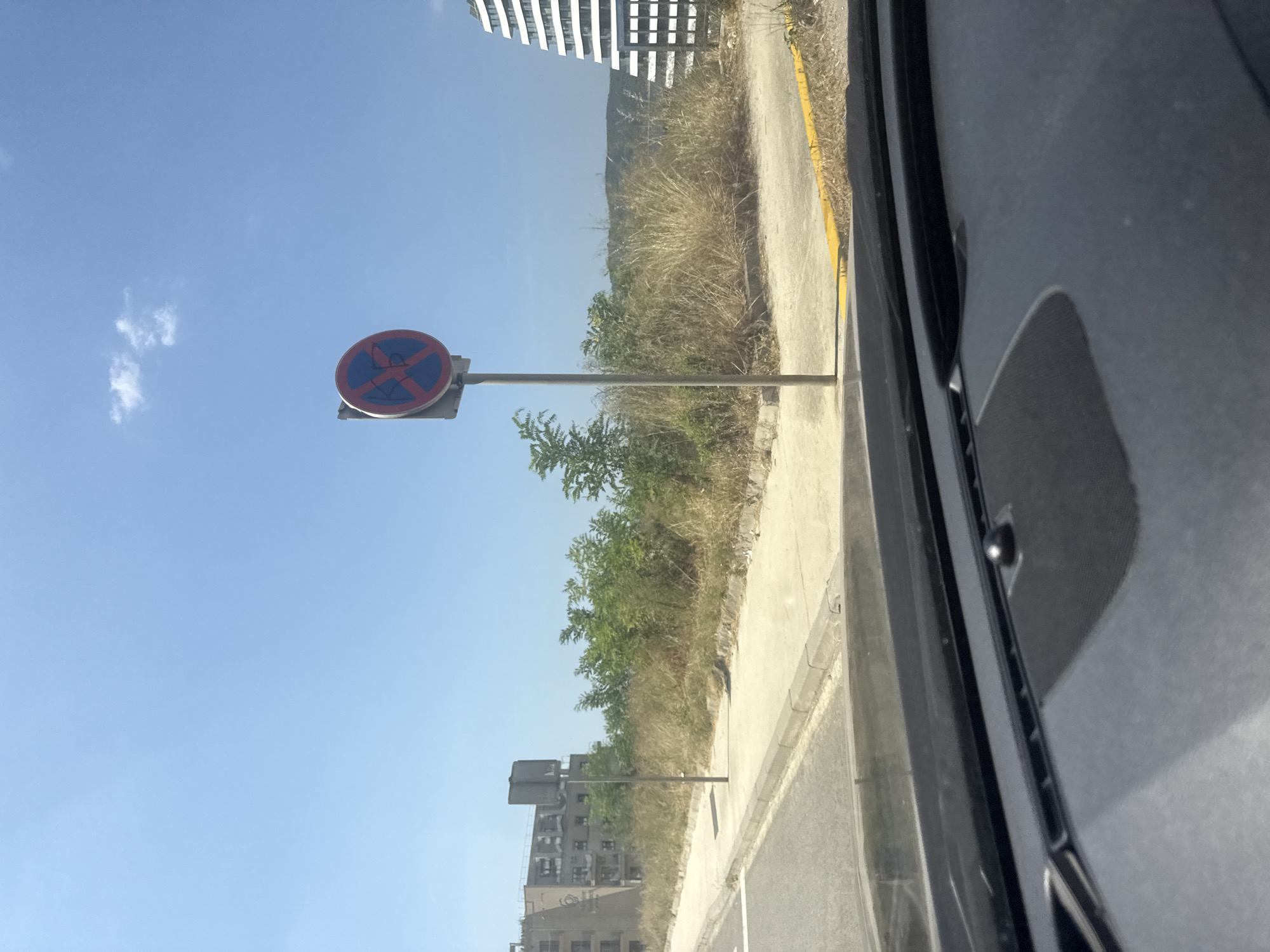 The image, taken from inside a vehicle, shows a street scene in Boreti, Montenegro. In the foreground, the vehicle's dashboard is visible. A no parking sign with a red circle and a blue inner circle with a red cross is prominently displayed on a pole to the left of the frame. Beyond the sign, there is a paved walkway and a patch of dry grass and bushes. In the background, an unfinished building with several floors is seen on the left, and a modern, white, multi-story building with a curved facade is visible on the right. The sky is clear and blue with a few scattered clouds. The overall environment suggests a sunny day in a developing or partially constructed area.