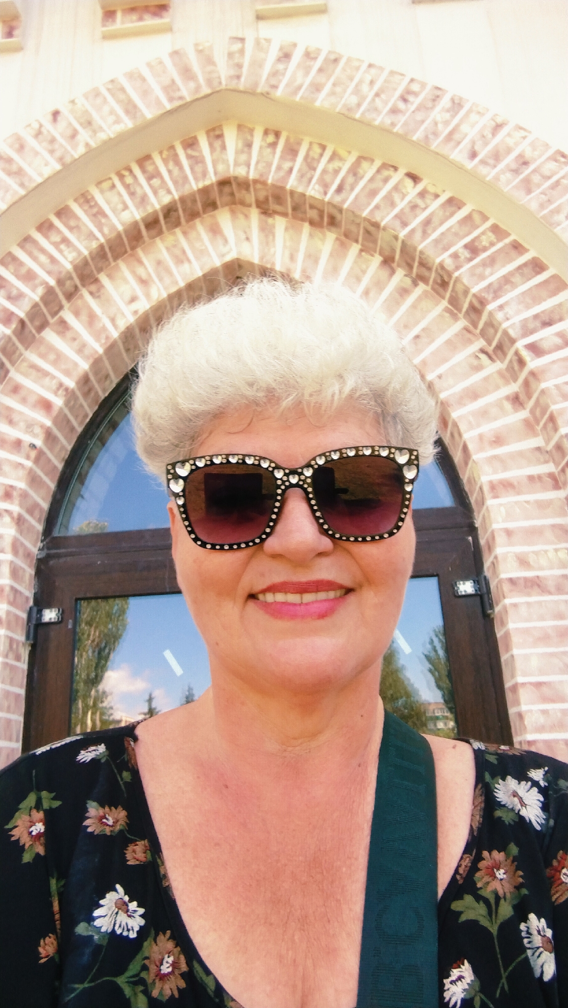 A medium shot captures a woman in her late 50s or early 60s, with short, permed white hair and bright pink lipstick, posing for a selfie. She is wearing large, decorative sunglasses with rhinestones and a black floral-print dress. A dark green strap is visible across her chest. The background features a brick archway and a glass door reflecting trees and the sky, suggesting an outdoor setting. This scene is located in the city of Horishni Plavni, Ukraine, specifically in front of the Ministry of Labour and Social Policy building, indicated by a sign on the door.