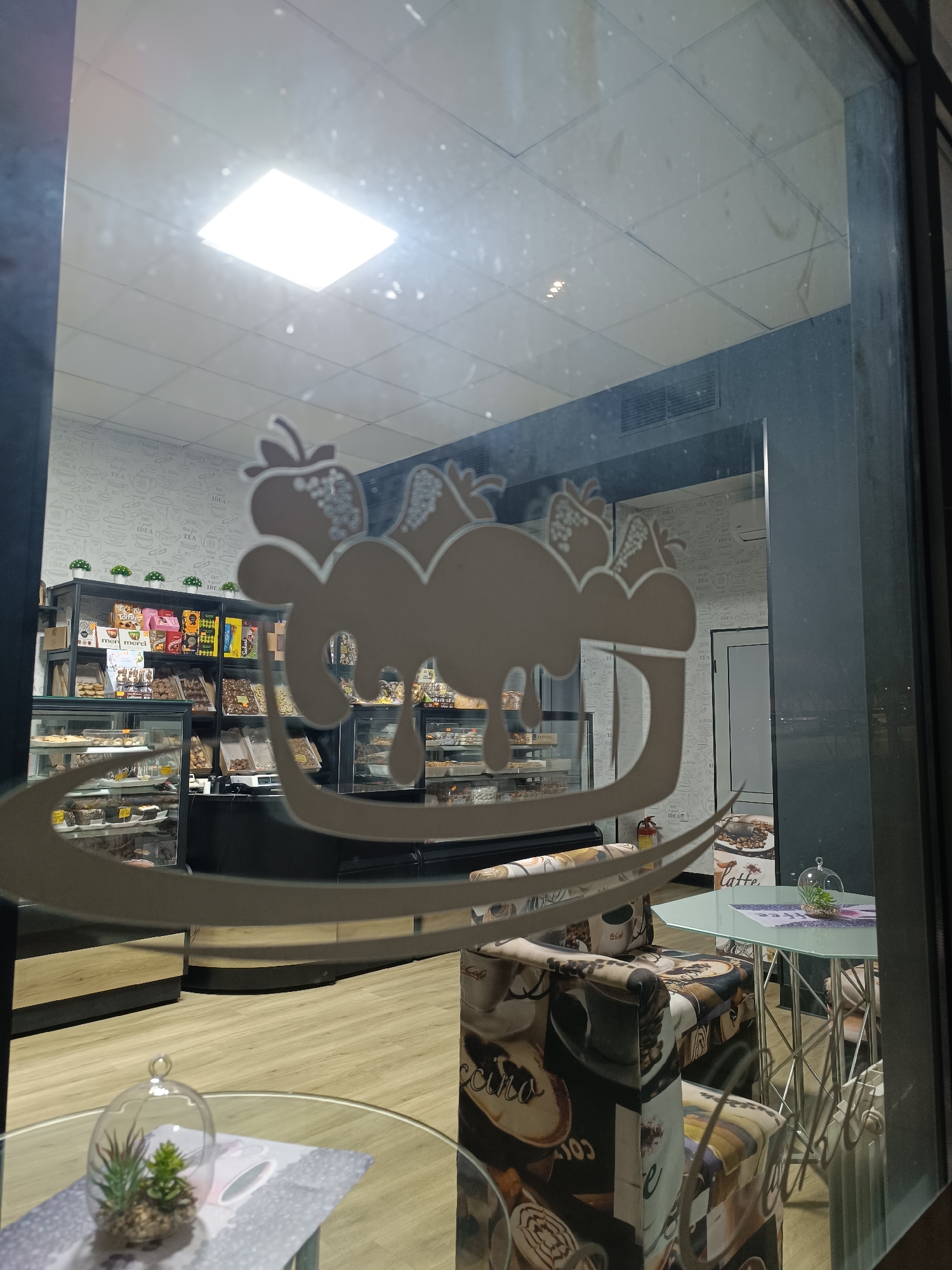 A view through the glass door of a shop in Pleven, Bulgaria, shows the interior of a cafe or bakery. The door has a white logo depicting a cloud-like shape with strawberries on top and a stylized drip or swirl design below. Inside, shelves and display cases are filled with various food items, including pastries and snacks. A patterned chair is placed in front of a small table with a glass top, on which sits a decorative terrarium. The walls inside have a patterned wallpaper with tea-related designs. The overall impression is of a modern, inviting bakery or cafe.